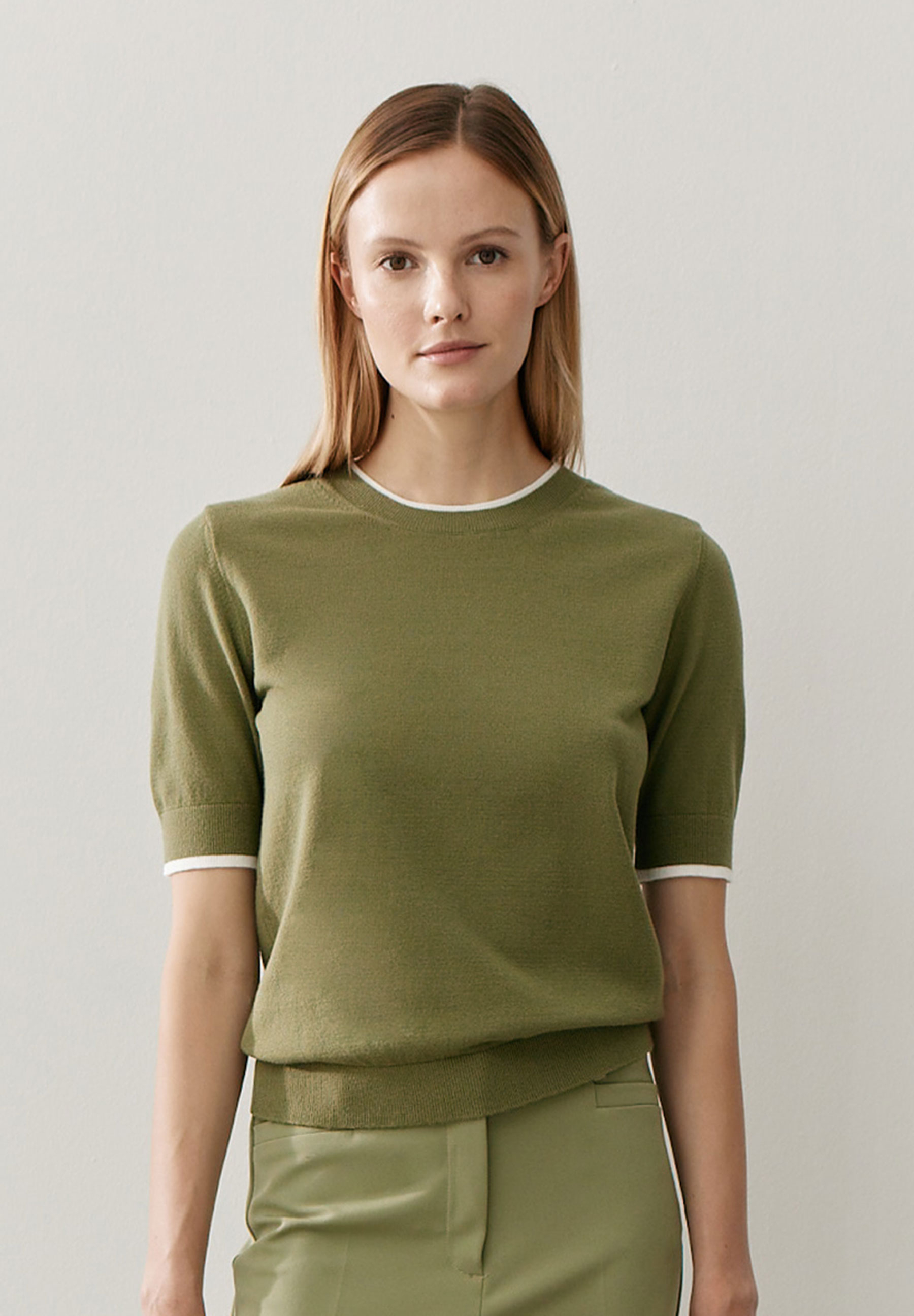 Eine Frau mit einem olivgrünen Feinstrick-Oberteil von MORE & MORE. Der Pullover hat weiße Akzente an Ärmeln und Kragen.