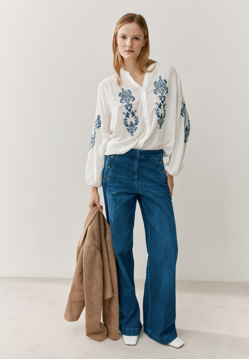 Eine Frau präsentiert eine weiße MORE & MORE Bluse mit blauen Stickereien und weite Jeans. Eine Frau präsentiert eine weiße MORE & MORE Bluse mit blauen Stickereien und weite Jeans.