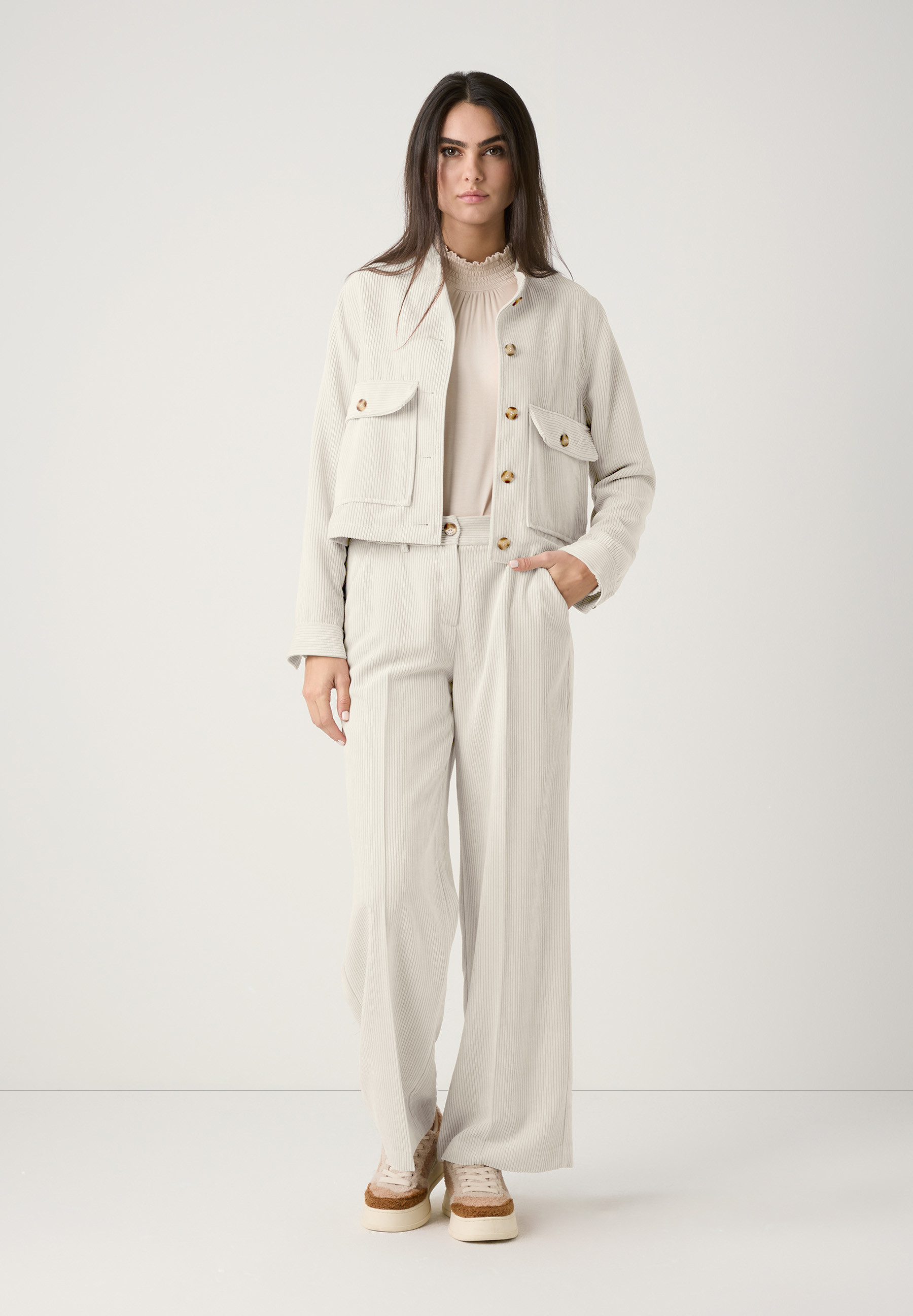 Eine Frau in einem MORE & MORE Cord-Ensemble, bestehend aus Oberteil und Hose, für angenehmen Tragekomfort und einen stilvollen Look. Eine Frau in einem MORE & MORE Cord-Ensemble, bestehend aus Oberteil und Hose, für angenehmen Tragekomfort und einen stilvollen Look.