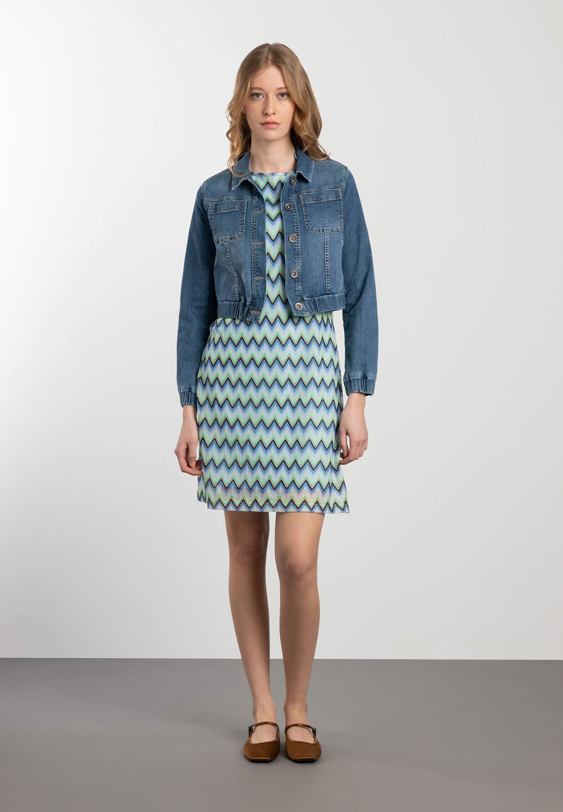 Eine Frau trägt eine MORE & MORE Jeansjacke über einem blau-grünen Kleid mit Zickzackmuster. Eine Frau trägt eine MORE & MORE Jeansjacke über einem blau-grünen Kleid mit Zickzackmuster.