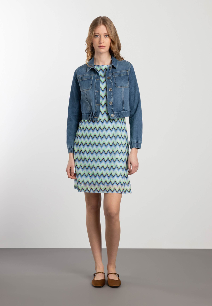 Eine Frau trägt eine MORE & MORE Jeansjacke über einem blau-grünen Kleid mit Zickzackmuster. Eine Frau trägt eine MORE & MORE Jeansjacke über einem blau-grünen Kleid mit Zickzackmuster.