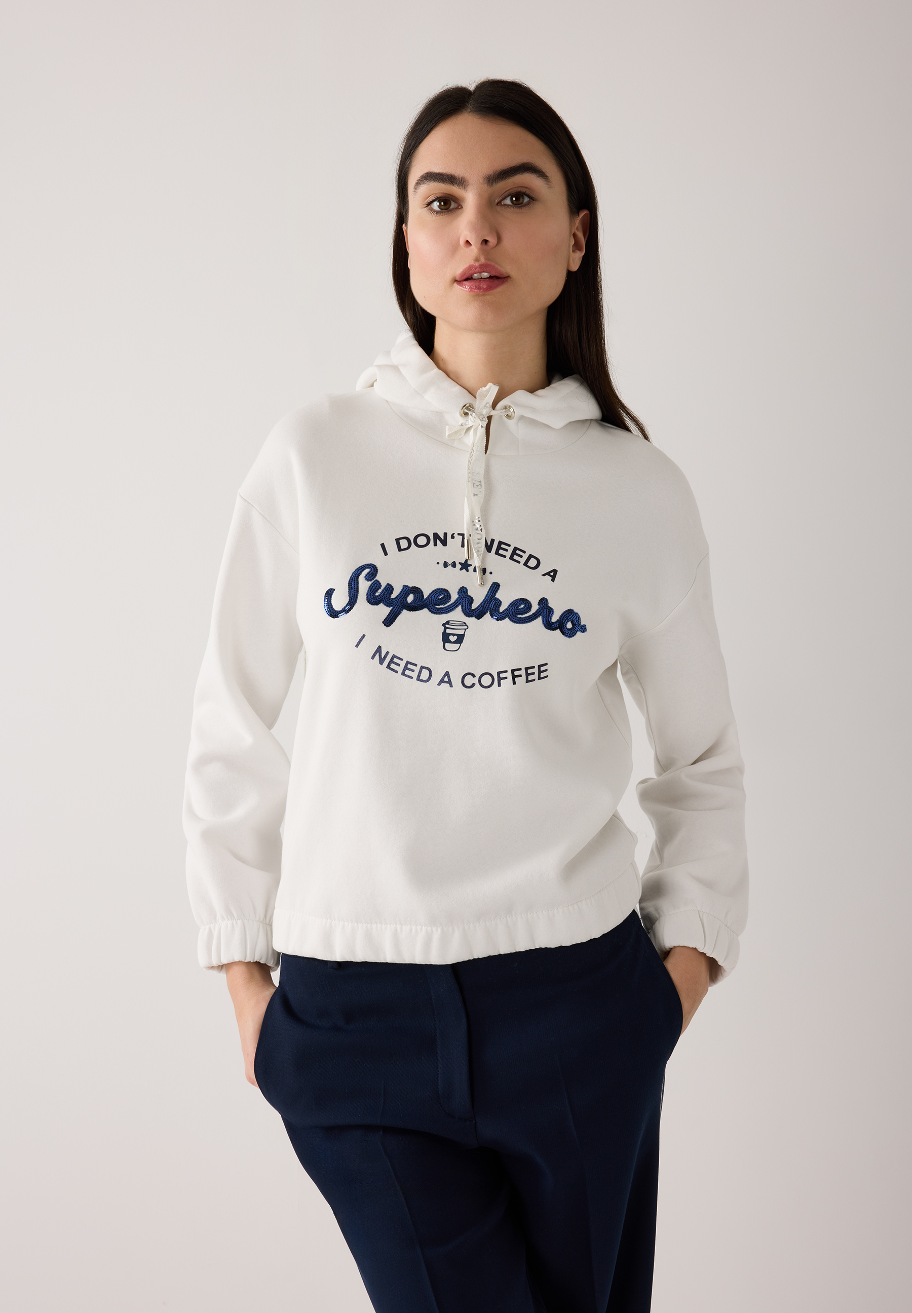 Eine Frau trägt ein MORE & MORE Sweatshirt mit der Aufschrift: 'I don't need a Superhero, I need a coffee'.