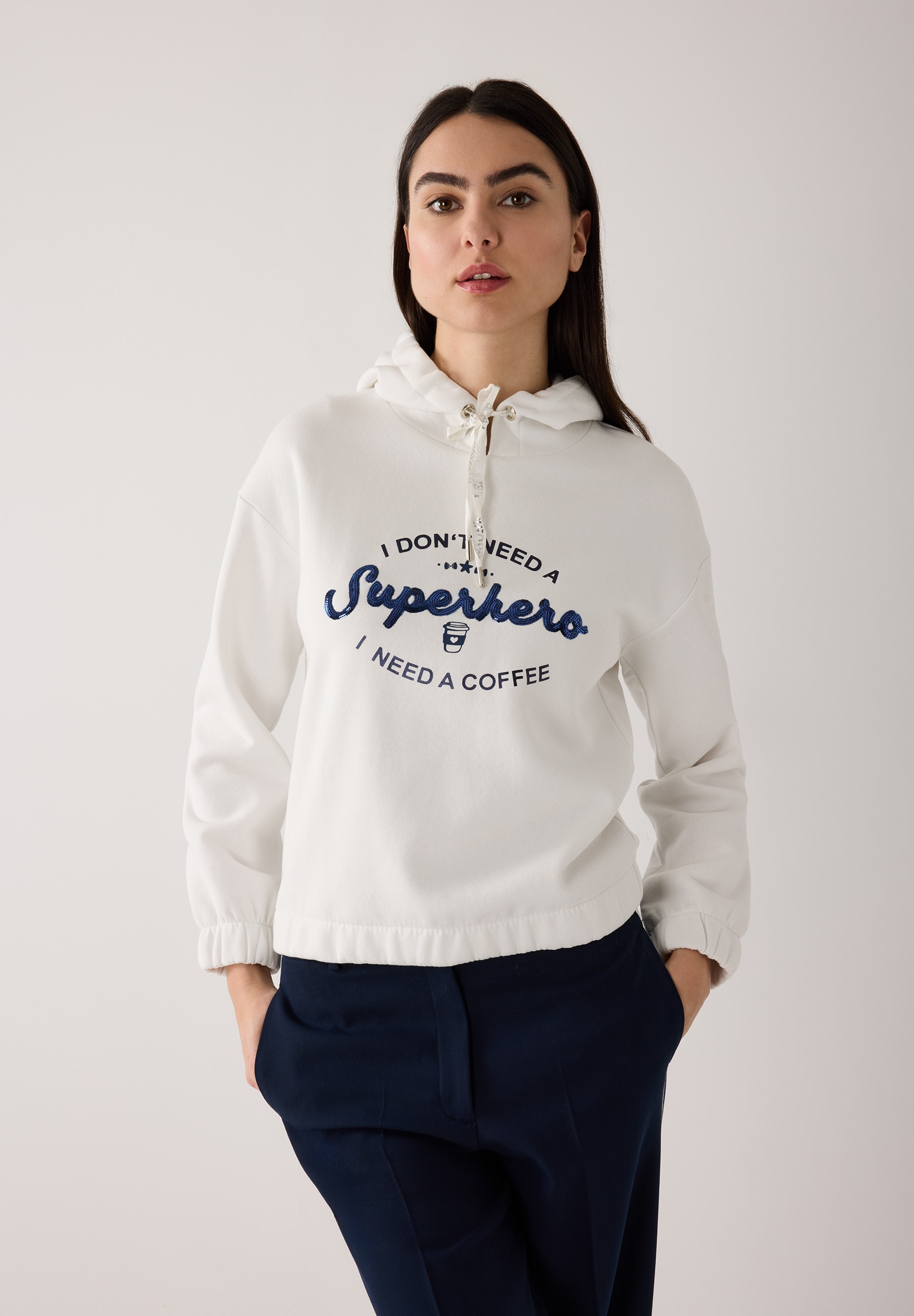 Eine Frau trägt ein MORE & MORE Sweatshirt mit der Aufschrift: 'I don't need a Superhero, I need a coffee'.
