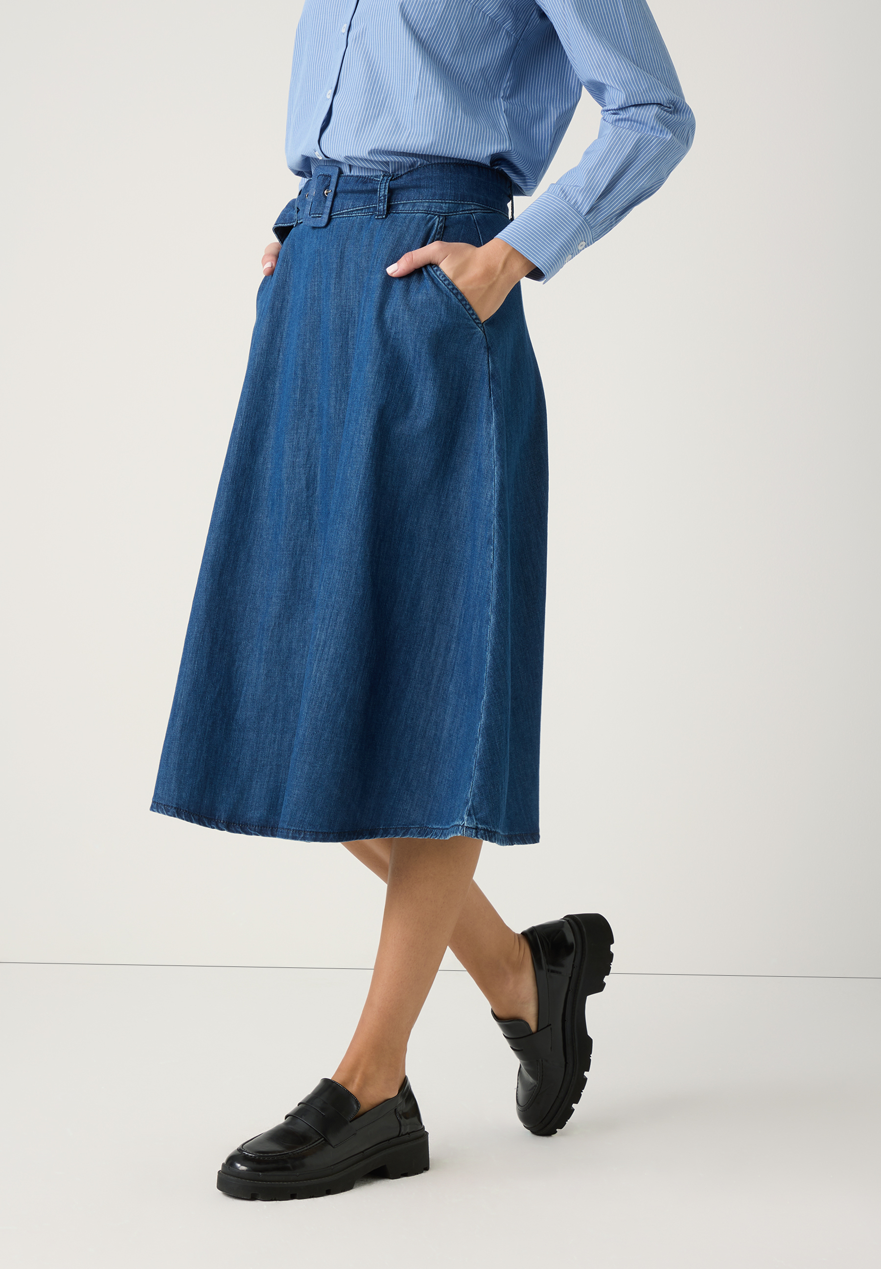 Ein Model präsentiert einen Jeansrock von MORE & MORE mit angenehmem Tragekomfort, kombiniert mit einer blau gestreiften Bluse und schwarzen Loafers.