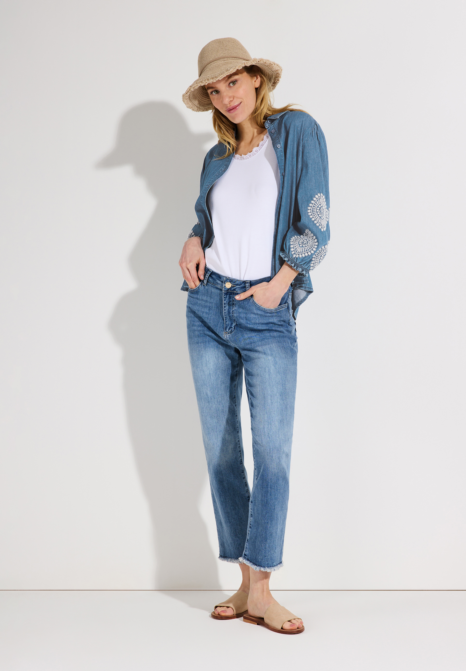 Eine Frau mit Hut und MORE & MORE Jeansbluse mit Stickerei steht locker da. Eine Frau mit Hut und MORE & MORE Jeansbluse mit Stickerei steht locker da.