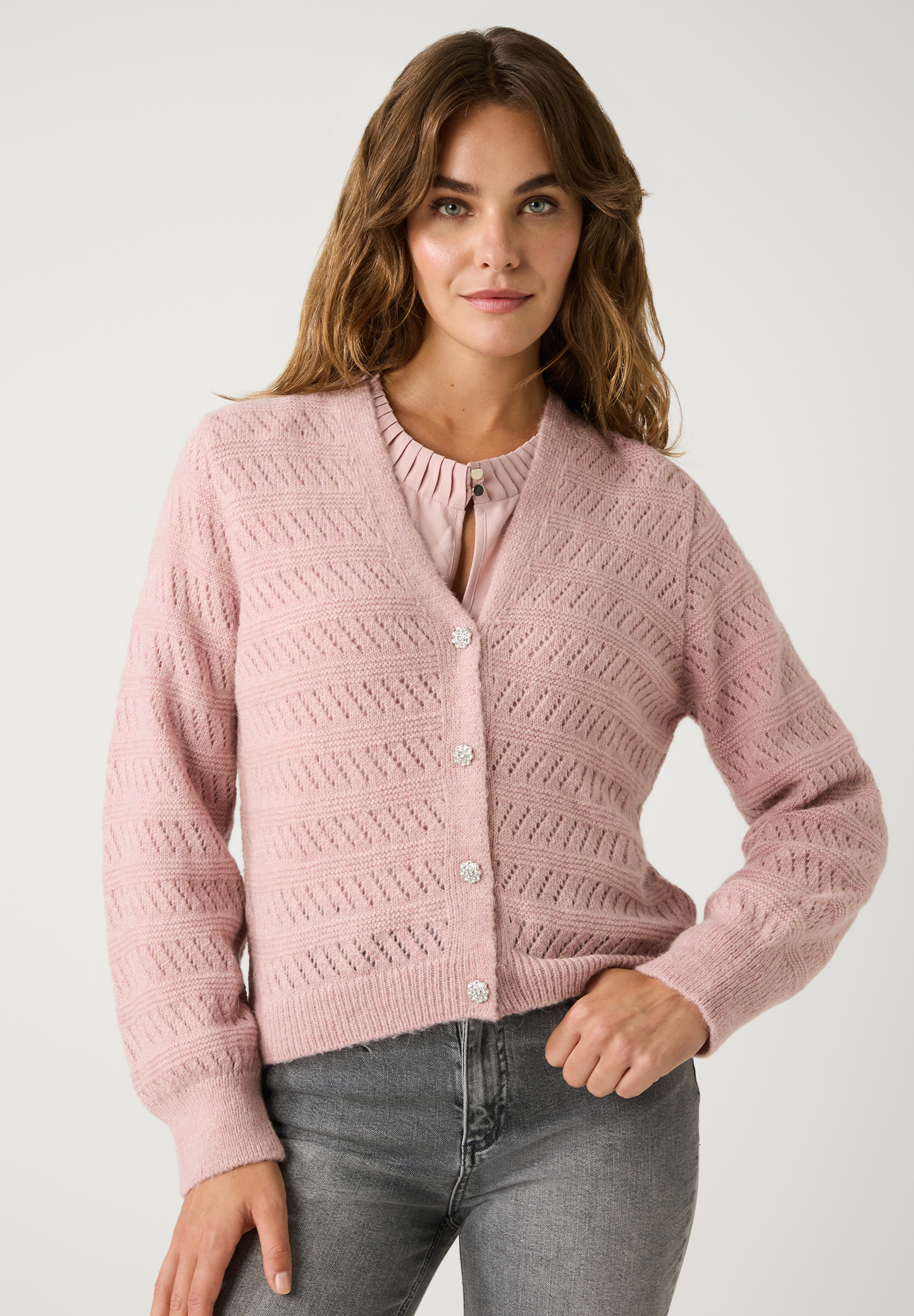 Eine Frau trägt einen rosa Lochstrick-Cardigan der Marke MORE & MORE mit glitzernden Knöpfen und eine graue Jeans.