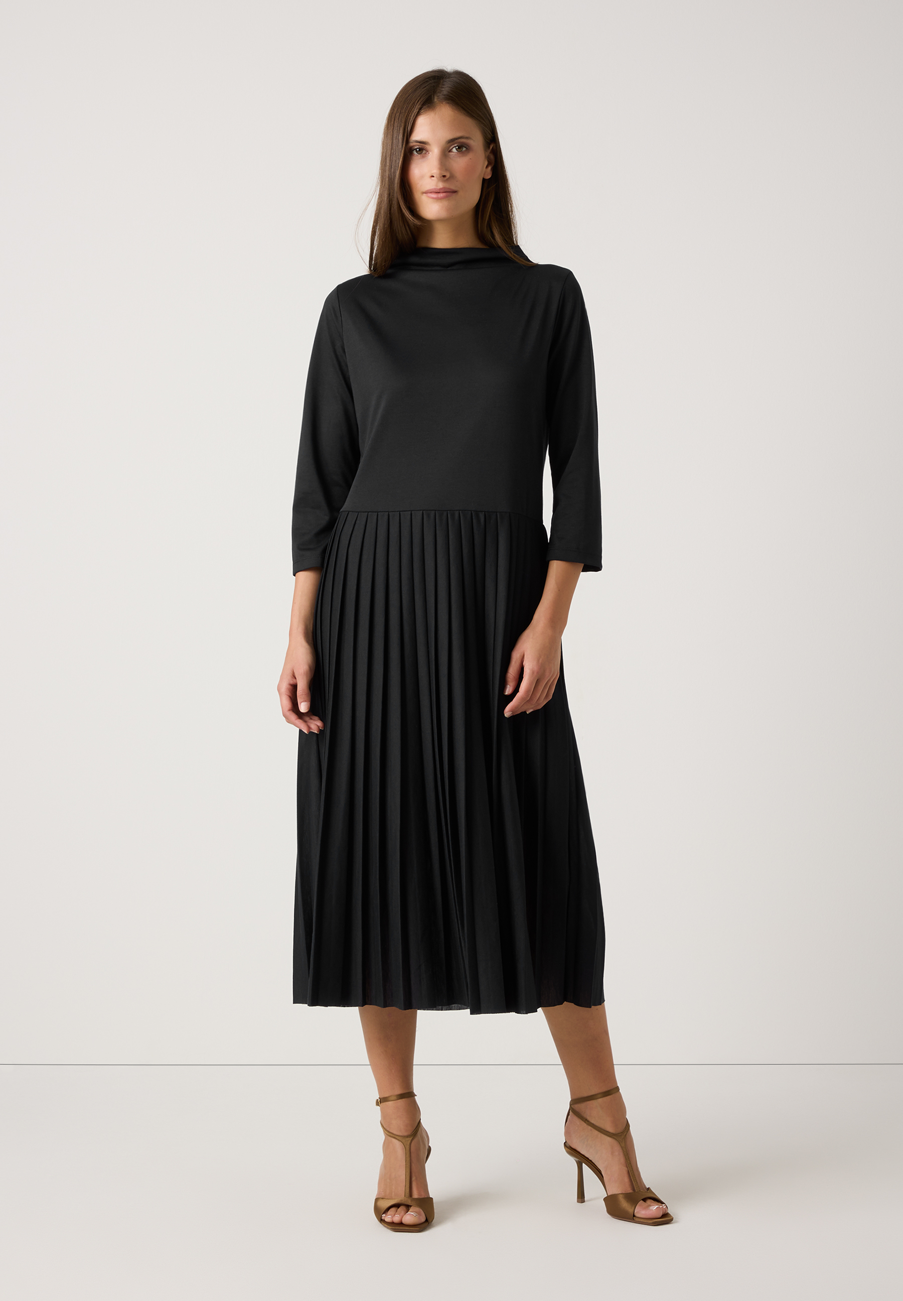 Eine Frau in einem schwarzen Jersey-Oberteil/Kleid von MORE & MORE mit 3/4-Ärmeln und Faltenrock. Dazu trägt sie goldene Riemchen-Sandaletten. Eine Frau in einem schwarzen Jersey-Oberteil/Kleid von MORE & MORE mit 3/4-Ärmeln und Faltenrock. Dazu trägt sie goldene Riemchen-Sandaletten.