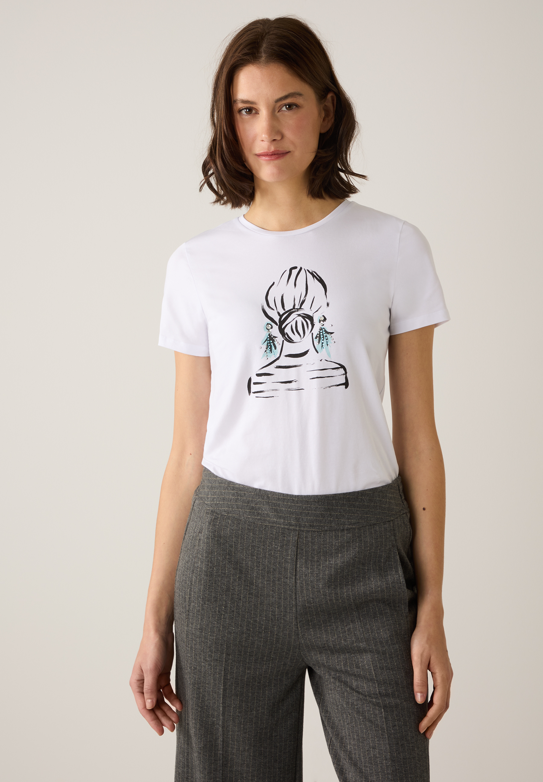 Eine Frau trägt ein weißes MORE & MORE T-Shirt mit Grafik und eine graue Stoffhose. Eine Frau trägt ein weißes MORE & MORE T-Shirt mit Grafik und eine graue Stoffhose.