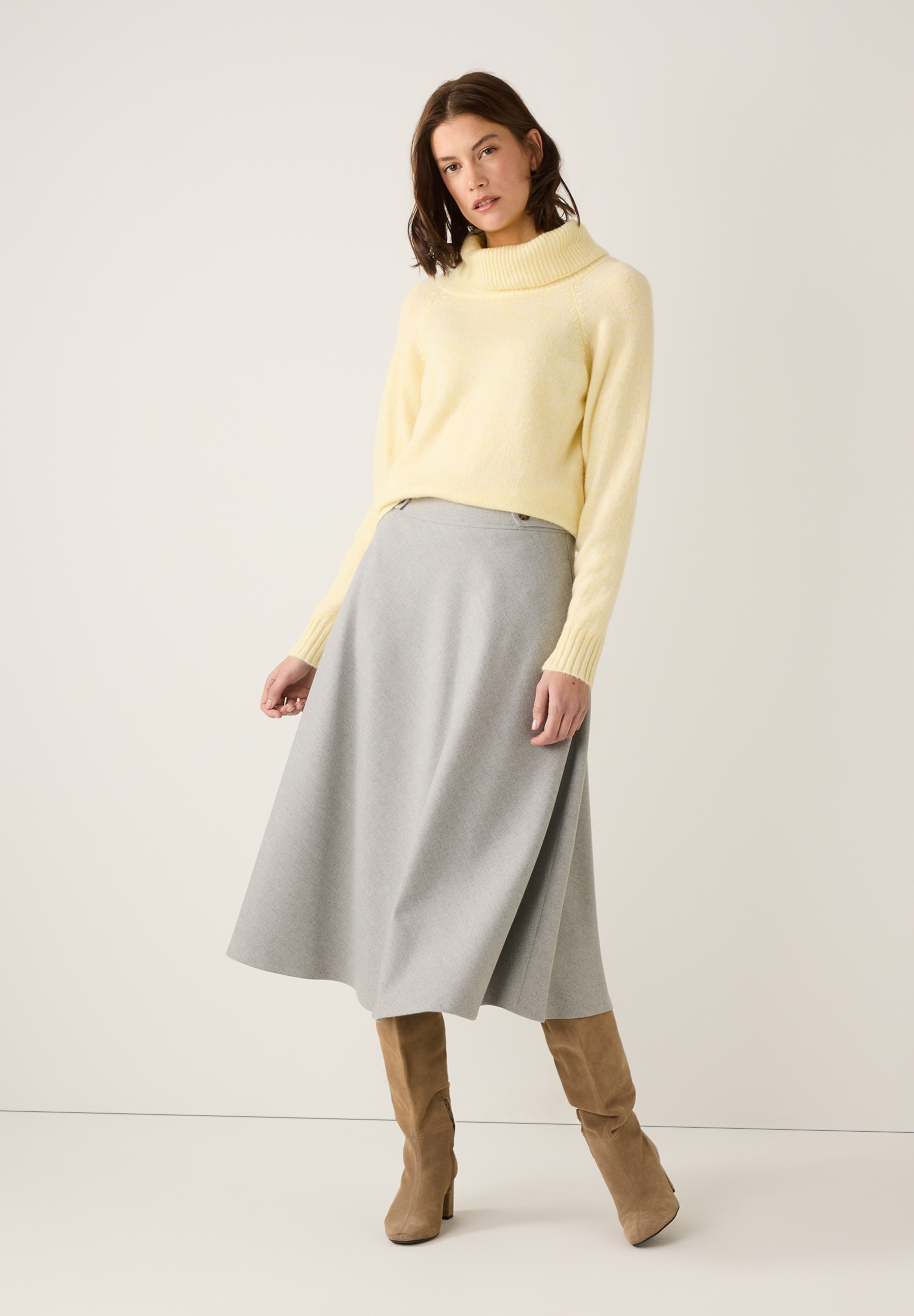 Eine Frau in MORE & MORE Strickmode: gelber Pullover, grauer Rock und braune Stiefel. Eine Frau in MORE & MORE Strickmode: gelber Pullover, grauer Rock und braune Stiefel.