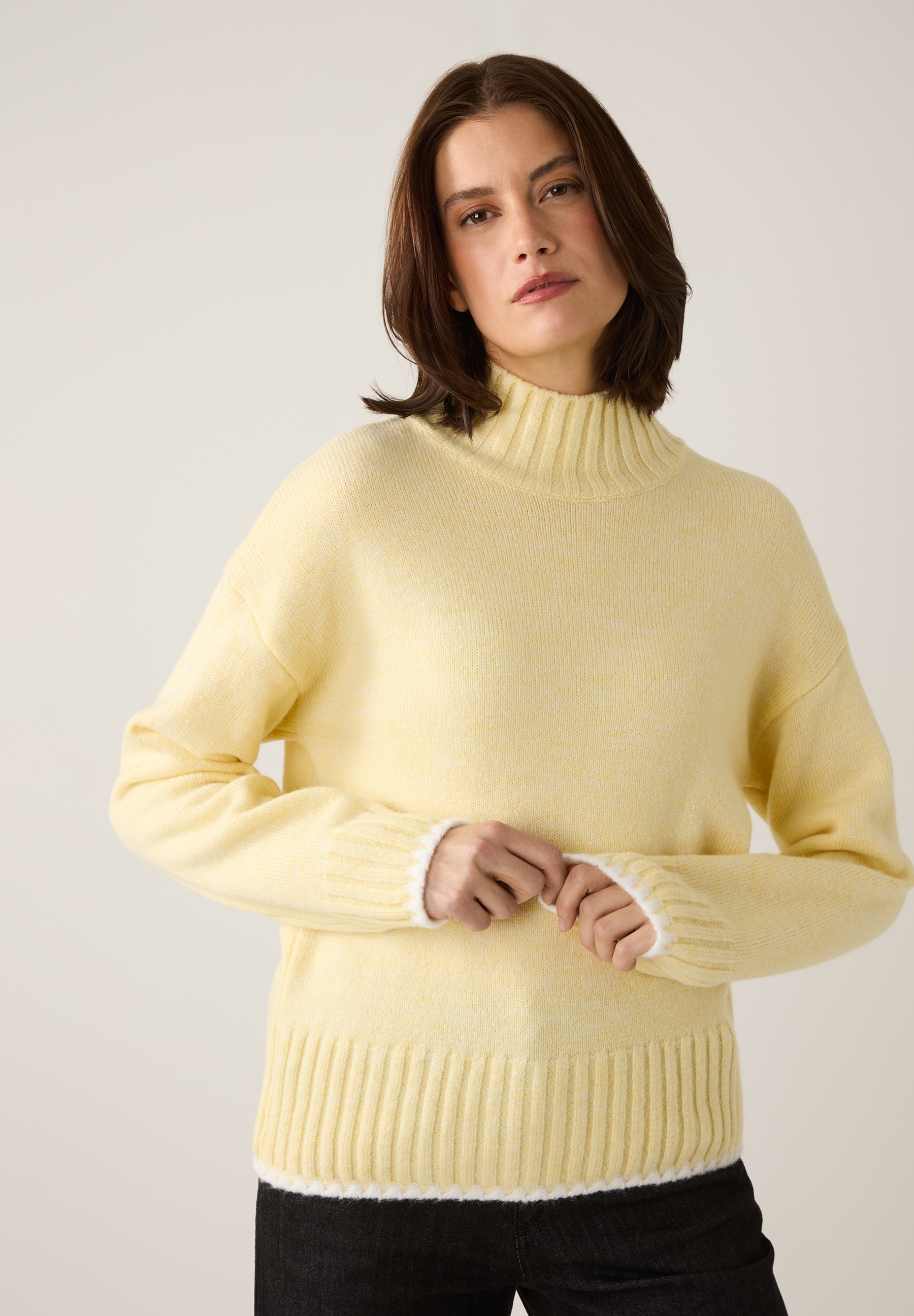 Eine Frau trägt einen gelben Strickpullover von MORE & MORE mit Rollkragen und weißen Details an Ärmeln und Saum.