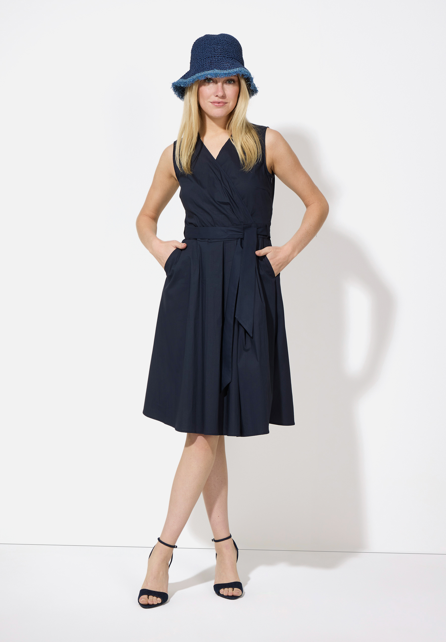 Eine Frau mit einem MORE & MORE Hut, einem ärmellosen Wickelkleid und Riemchensandalen posiert vor einem weißen Hintergrund.