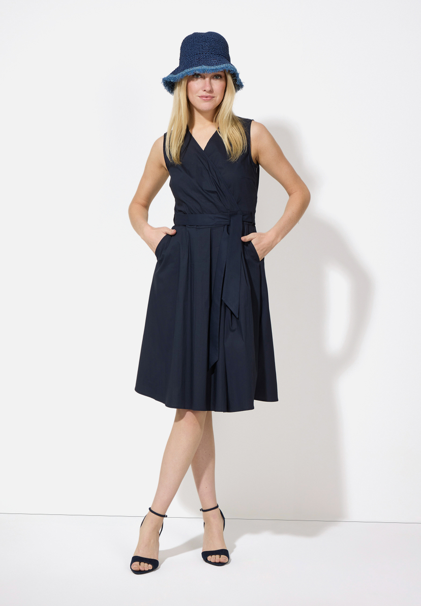Eine Frau mit einem MORE & MORE Hut, einem ärmellosen Wickelkleid und Riemchensandalen posiert vor einem weißen Hintergrund.