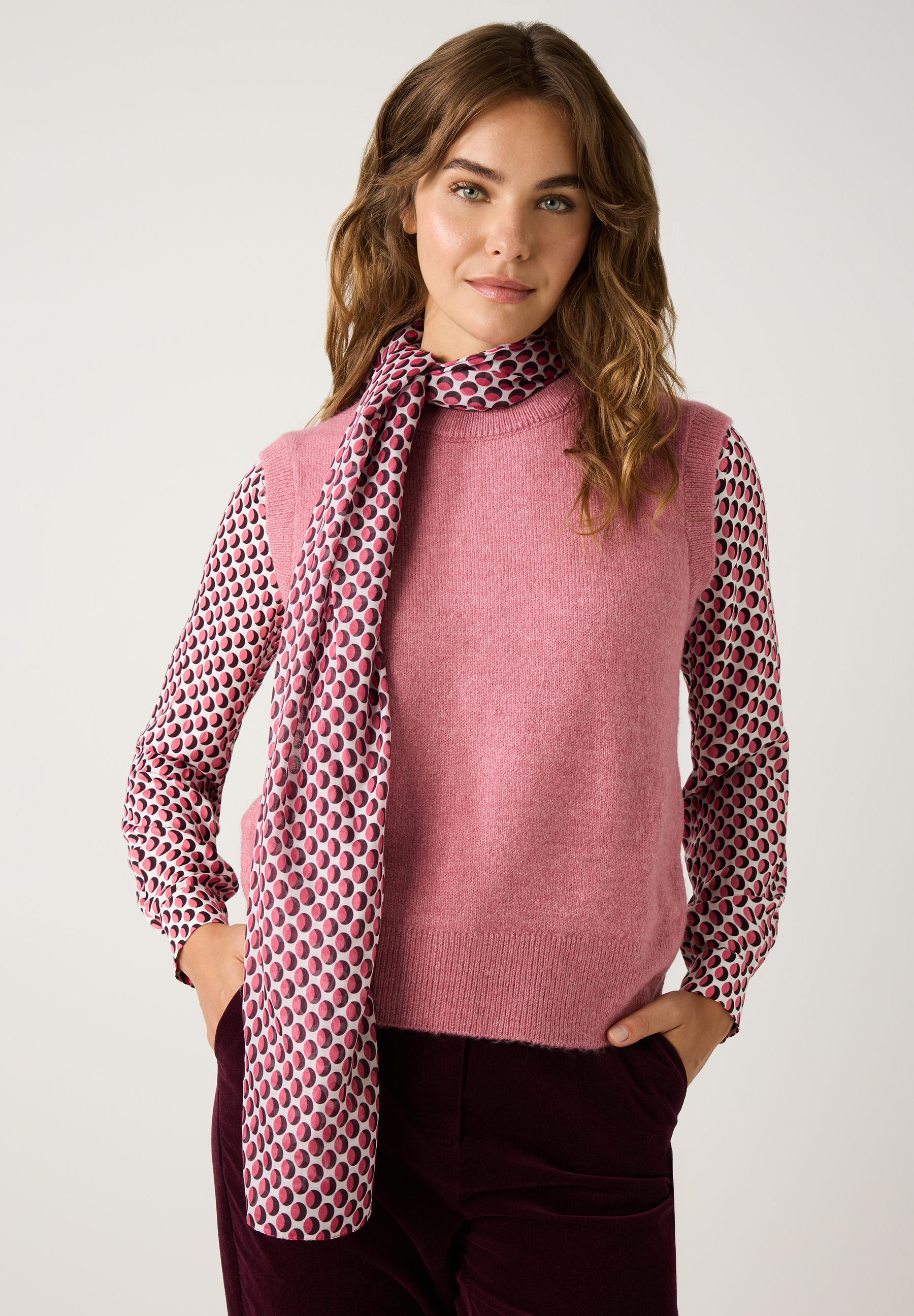 Eine Frau mit MORE & MORE Accessoires: Schal, Bluse, Pullunder in Rosa und Hose in Bordeaux vor einem weißen Hintergrund.