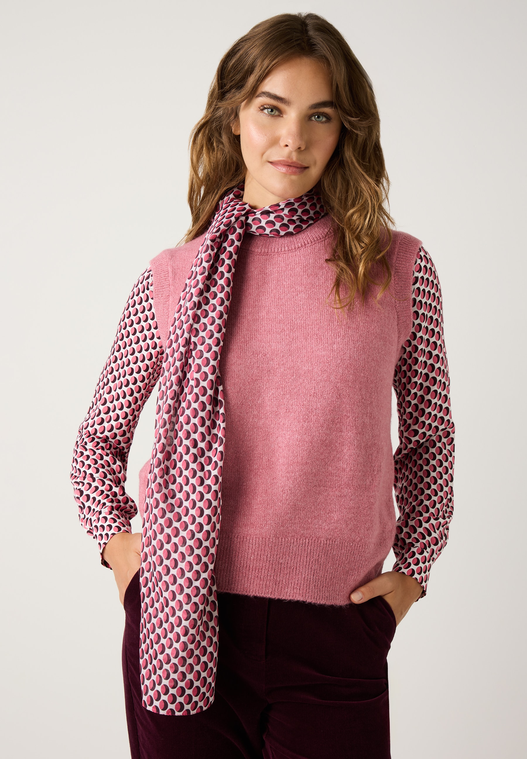 Eine Frau mit MORE & MORE Accessoires: Schal, Bluse, Pullunder in Rosa und Hose in Bordeaux vor einem weißen Hintergrund.