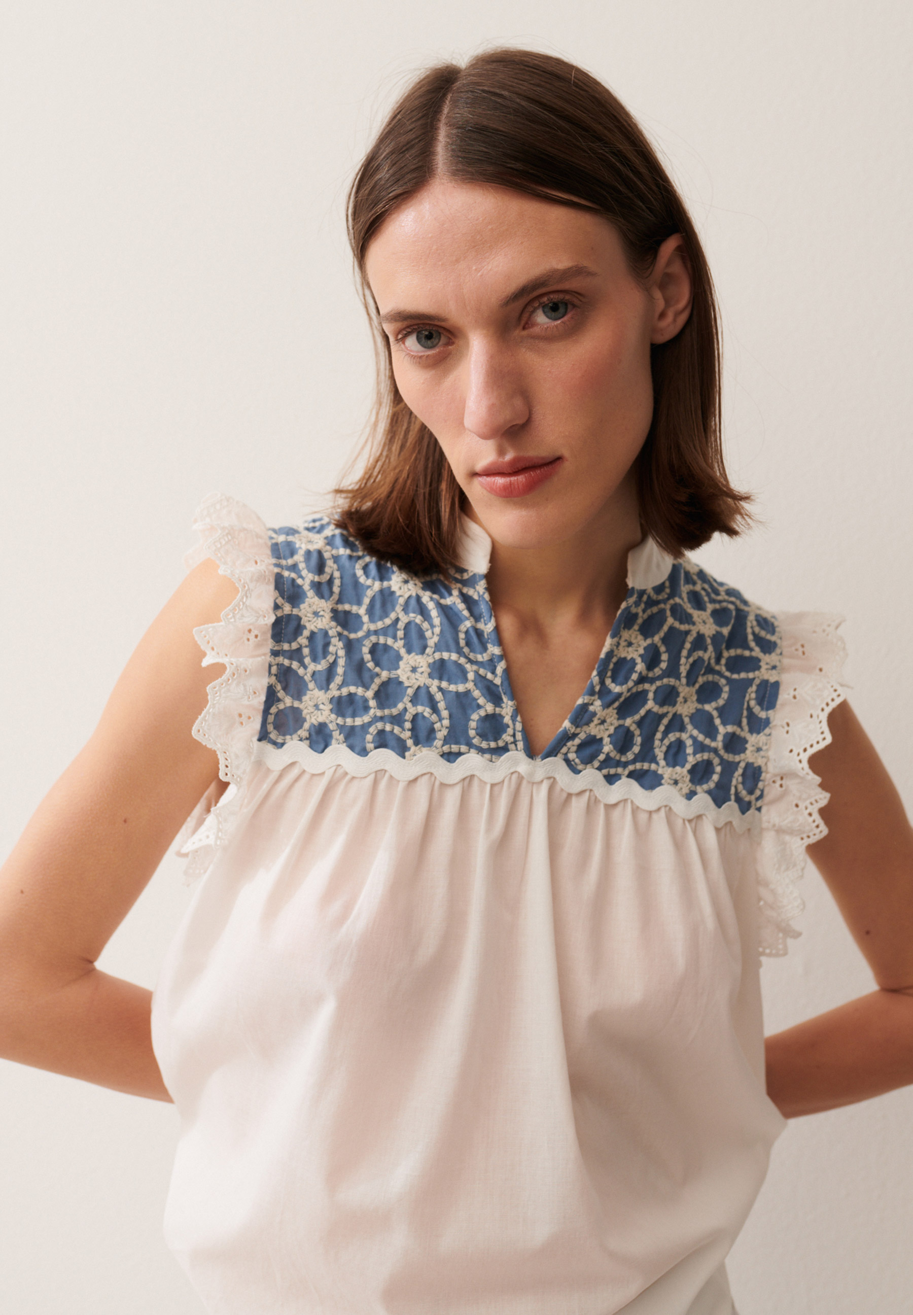 Eine Frau präsentiert eine weiße MORE & MORE Bluse mit blauen Stickereien und Spitzenärmeln vor einem hellen Hintergrund.