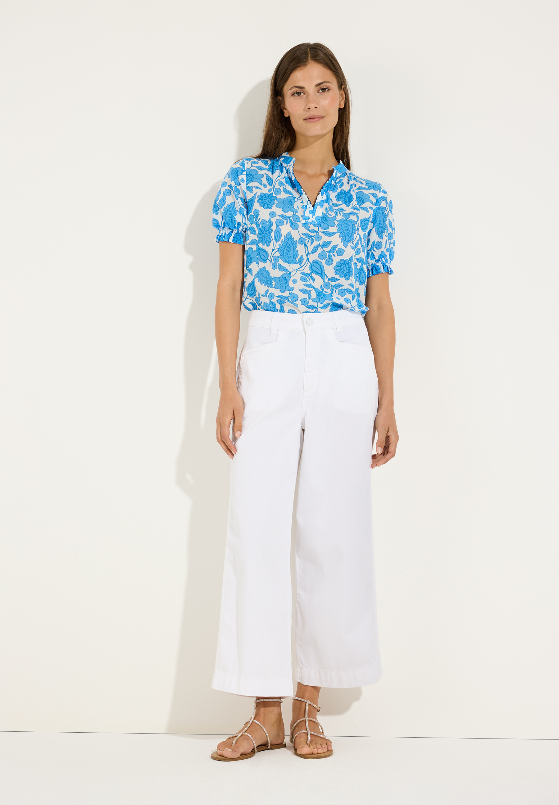 MORE & MORE Bluse: Eine stehende Frau trägt eine blaue Blumen-Bluse mit V-Ausschnitt, weiße Culottes und Sandalen. MORE & MORE Bluse: Eine stehende Frau trägt eine blaue Blumen-Bluse mit V-Ausschnitt, weiße Culottes und Sandalen.