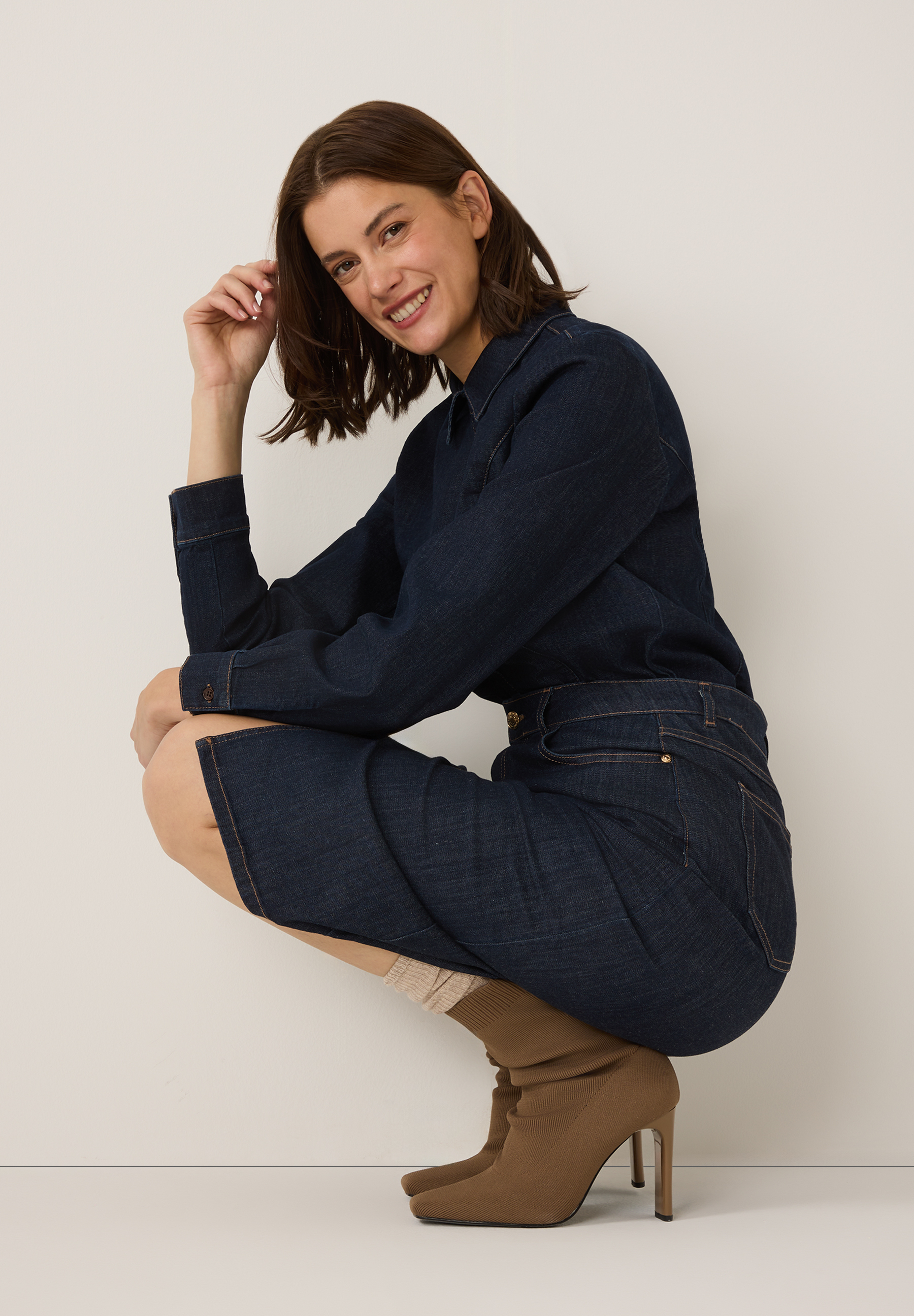 Eine lächelnde Frau hockt in einer MORE & MORE Jeansbluse und einem Rock mit braunen Stiefeln. Eine lächelnde Frau hockt in einer MORE & MORE Jeansbluse und einem Rock mit braunen Stiefeln.