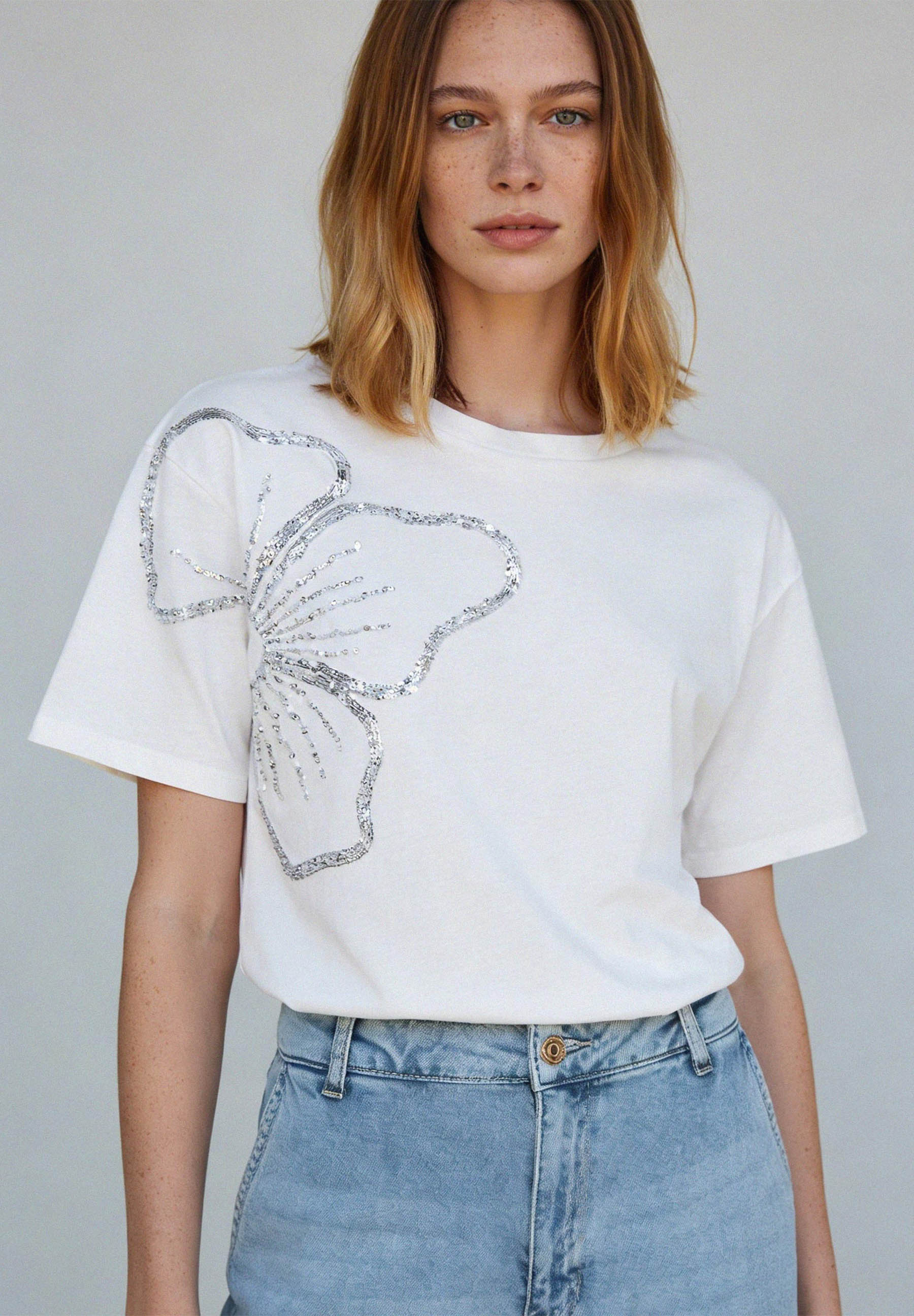 Eine Frau mit einem weißen T-Shirt von MORE & MORE mit silberner Blumenapplikation und hellblauen Jeans. Eine Frau mit einem weißen T-Shirt von MORE & MORE mit silberner Blumenapplikation und hellblauen Jeans.