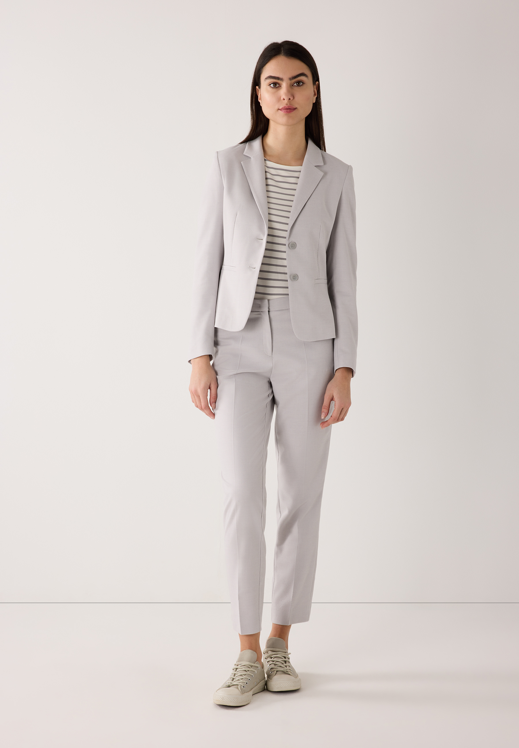 Eine Frau trägt ein graues MORE & MORE Blazer-Outfit mit einem gestreiften Oberteil und Sneakers vor einem weißen Hintergrund. Eine Frau trägt ein graues MORE & MORE Blazer-Outfit mit einem gestreiften Oberteil und Sneakers vor einem weißen Hintergrund.