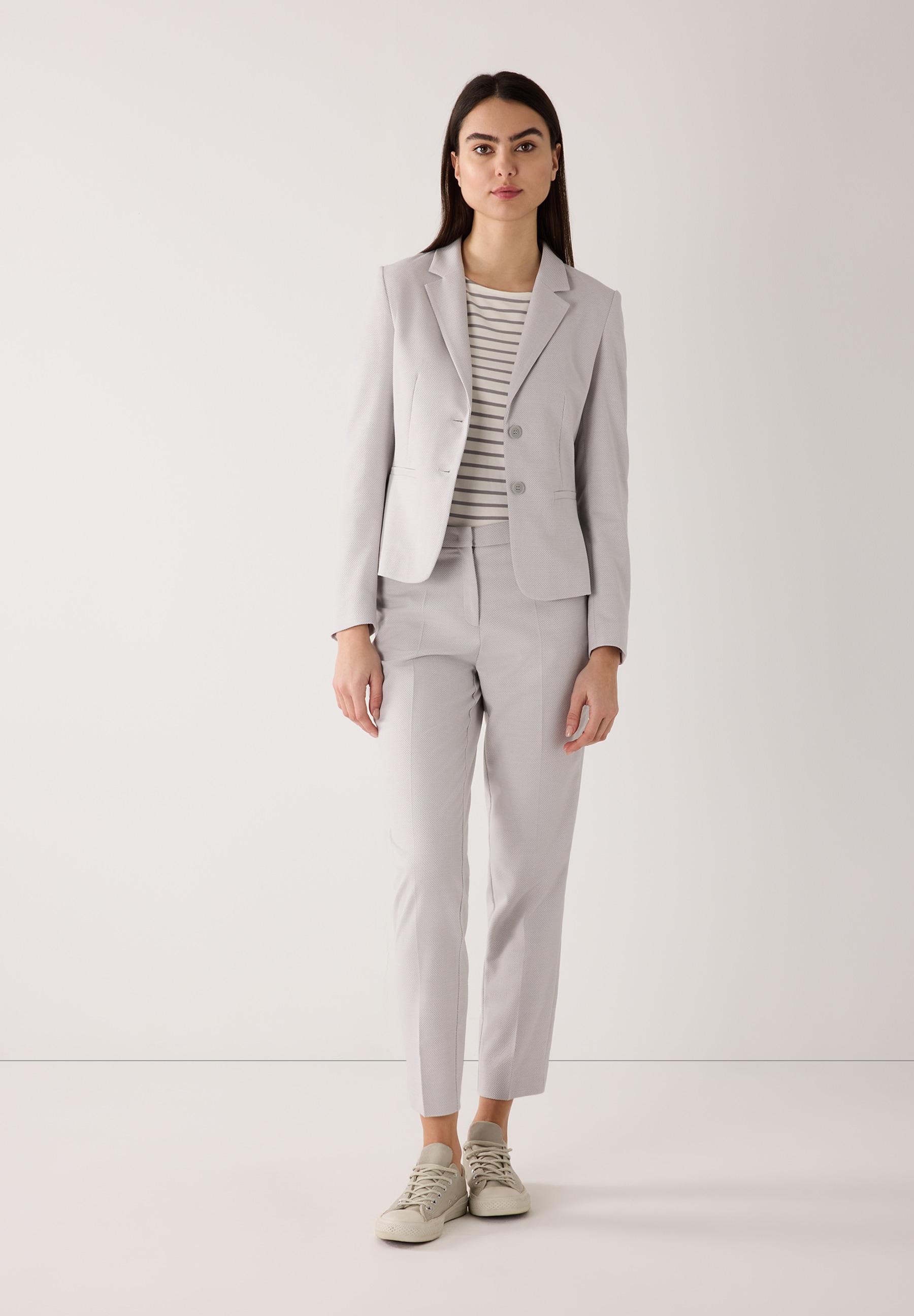 Eine Frau trägt ein graues MORE & MORE Blazer-Outfit mit einem gestreiften Oberteil und Sneakers vor einem weißen Hintergrund. Eine Frau trägt ein graues MORE & MORE Blazer-Outfit mit einem gestreiften Oberteil und Sneakers vor einem weißen Hintergrund.