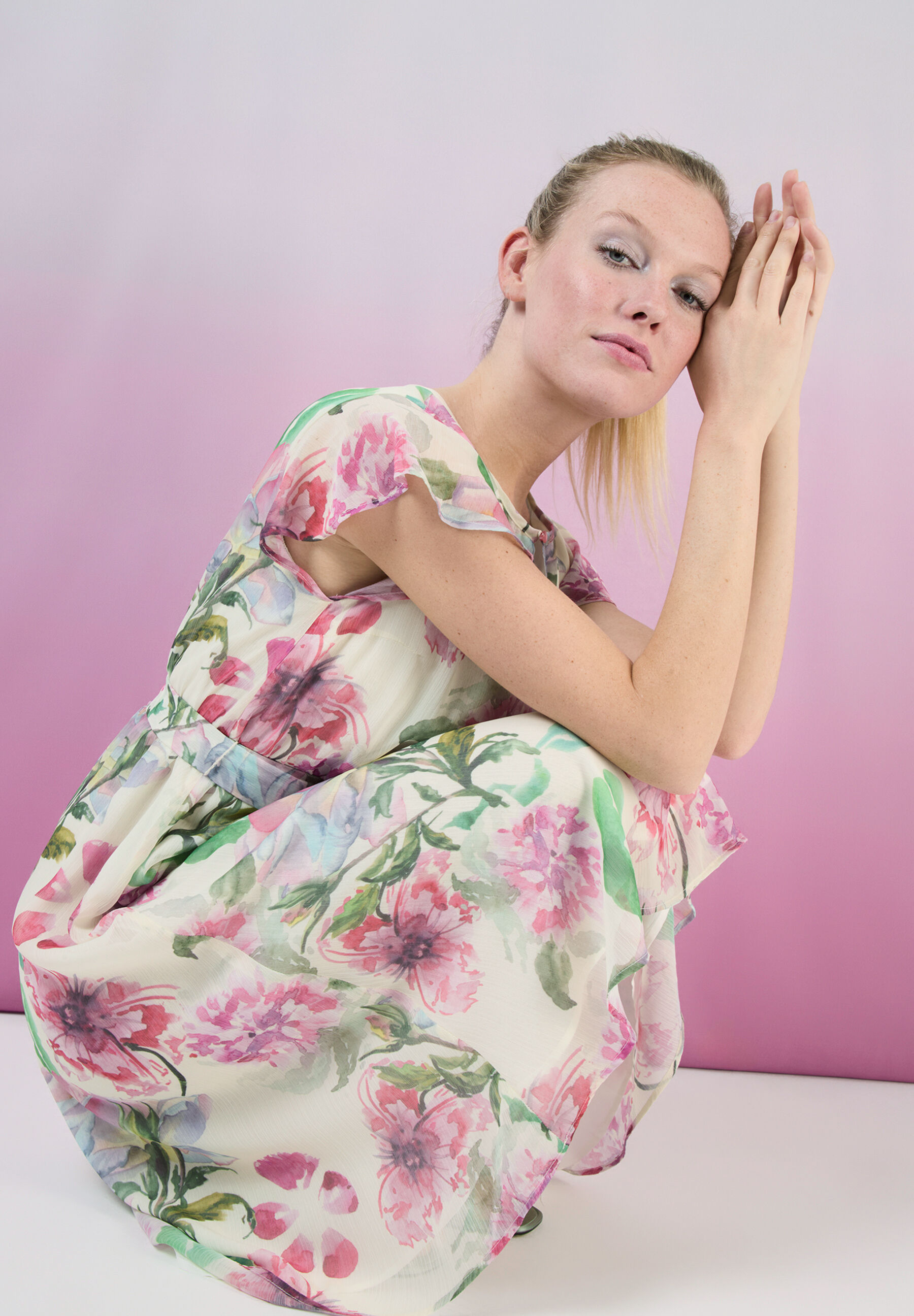 Eine Frau in einem Kleid mit Blumenmuster von MORE & MORE kniet vor einem rosa Hintergrund. Eine Frau in einem Kleid mit Blumenmuster von MORE & MORE kniet vor einem rosa Hintergrund.