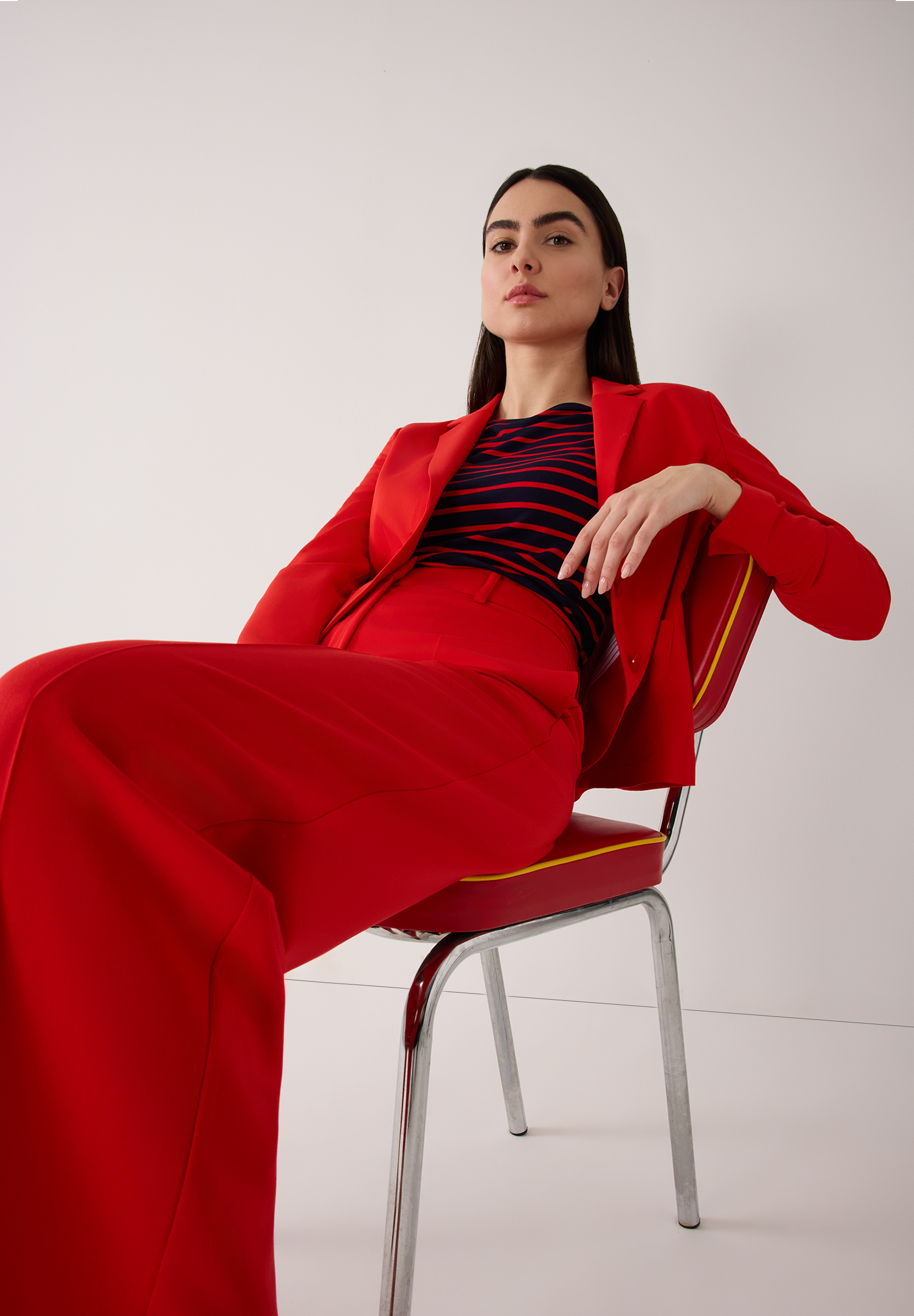 Eine Frau sitzt in rotem Hosenanzug von MORE & MORE auf einem Stuhl vor einem weißen Hintergrund. Eine Frau sitzt in rotem Hosenanzug von MORE & MORE auf einem Stuhl vor einem weißen Hintergrund.