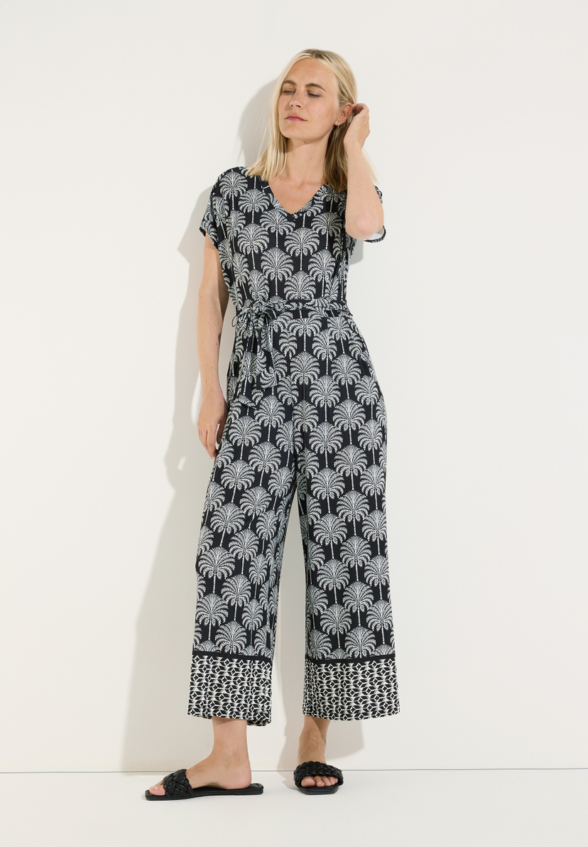 MORE & MORE Jersey-Hose im Palmenmuster. Eine Frau sitzt mit gekreuzten Beinen auf dem Boden und trägt eine schwarze Hose mit weißen Palmen- und geometrischen Mustern. Sie trägt schwarze geflochtene Sandalen. MORE & MORE Jersey-Hose im Palmenmuster. Eine Frau sitzt mit gekreuzten Beinen auf dem Boden und trägt eine schwarze Hose mit weißen Palmen- und geometrischen Mustern. Sie trägt schwarze geflochtene Sandalen.