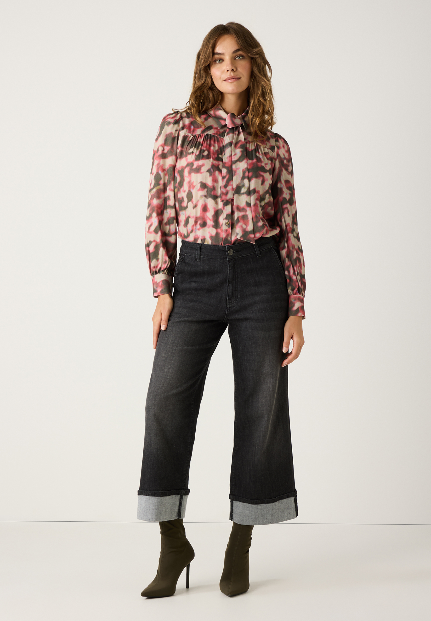 Eine Frau in MORE & MORE Bluse mit Muster, dunkler Jeans und Stiefeletten vor einem weißen Hintergrund. Eine Frau in MORE & MORE Bluse mit Muster, dunkler Jeans und Stiefeletten vor einem weißen Hintergrund.