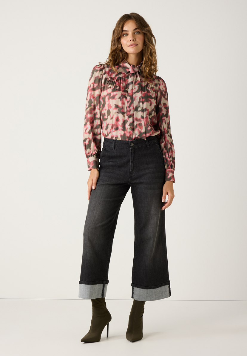 Eine Frau in MORE & MORE Bluse mit Muster, dunkler Jeans und Stiefeletten vor einem weißen Hintergrund. Eine Frau in MORE & MORE Bluse mit Muster, dunkler Jeans und Stiefeletten vor einem weißen Hintergrund.