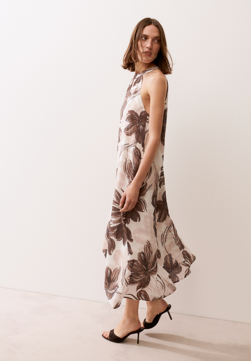 Eine Frau trägt ein MORE & MORE Kleid mit Blumenmuster und schwarze Sandalen mit Absatz vor einem hellen Hintergrund.