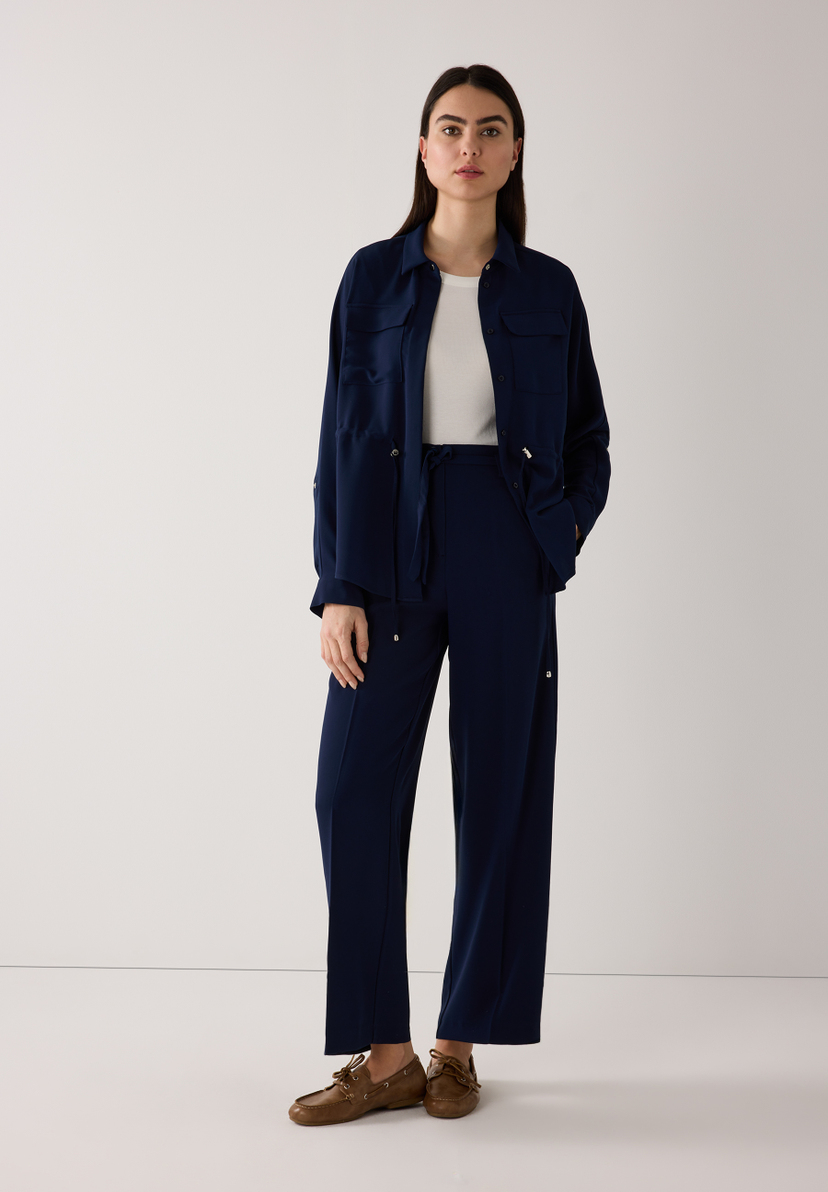 Eine Frau trägt ein marineblaues MORE & MORE Webware-Outfit, bestehend aus Bluse und Blazer, mit braunen Schuhen vor einem weißen Hintergrund. Eine Frau trägt ein marineblaues MORE & MORE Webware-Outfit, bestehend aus Bluse und Blazer, mit braunen Schuhen vor einem weißen Hintergrund.