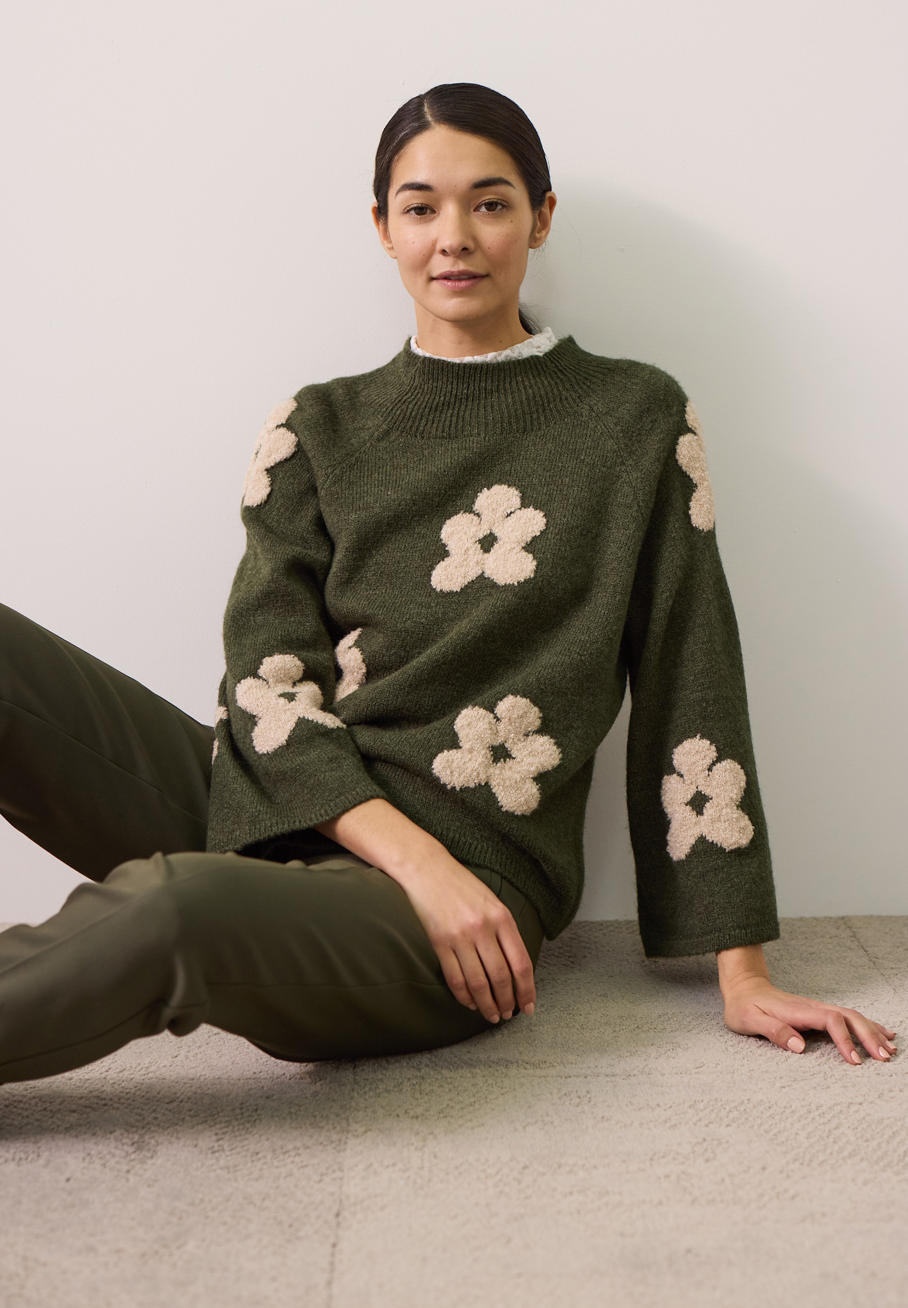 MORE & MORE Pullover: Eine Frau sitzt und trägt einen moosgrünen MORE & MORE Pullover mit großen, beigefarbenen Blumen.
