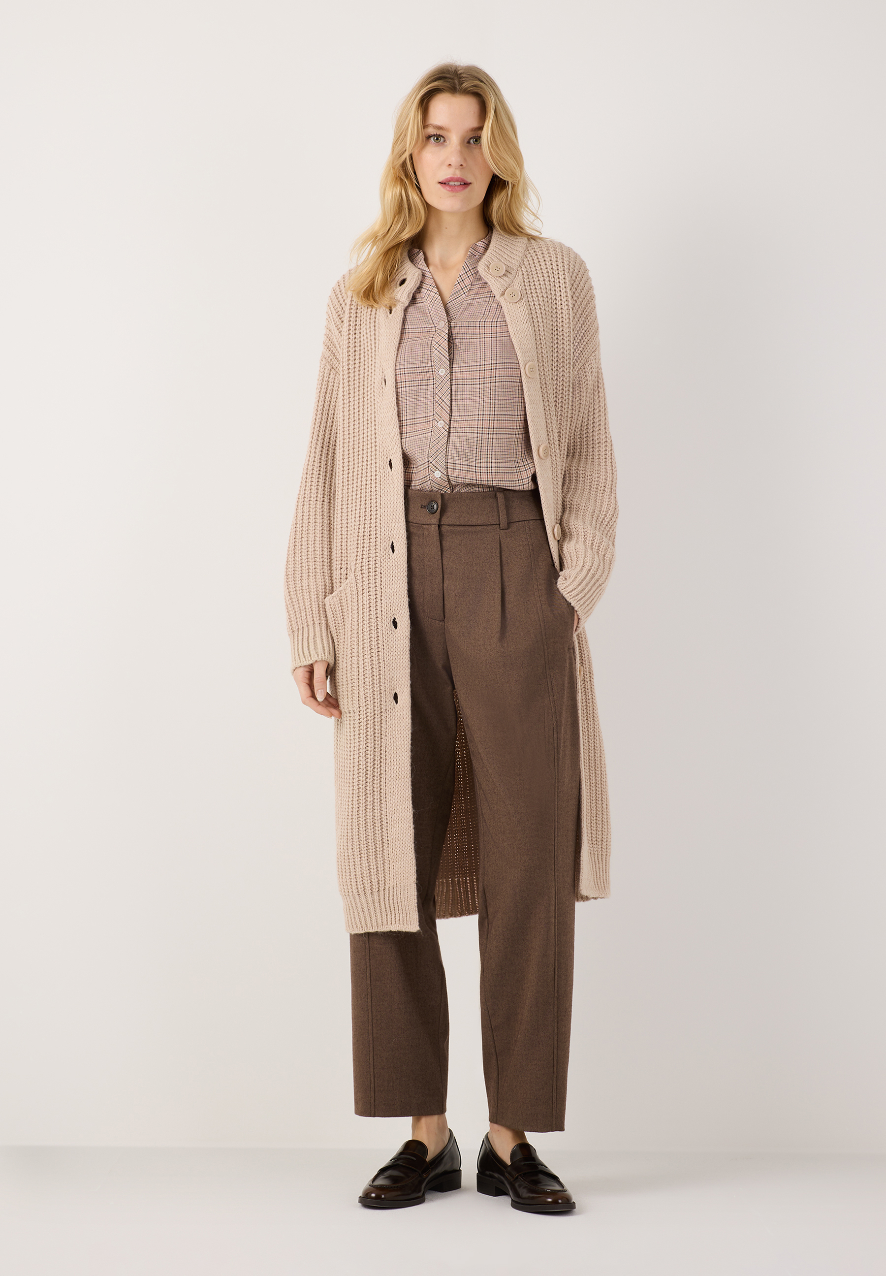 MORE & MORE Hose & Cardigan: Eine junge Frau trägt eine beigefarbene, locker gestrickte Strickjacke und eine braune MORE & MORE Jeans.