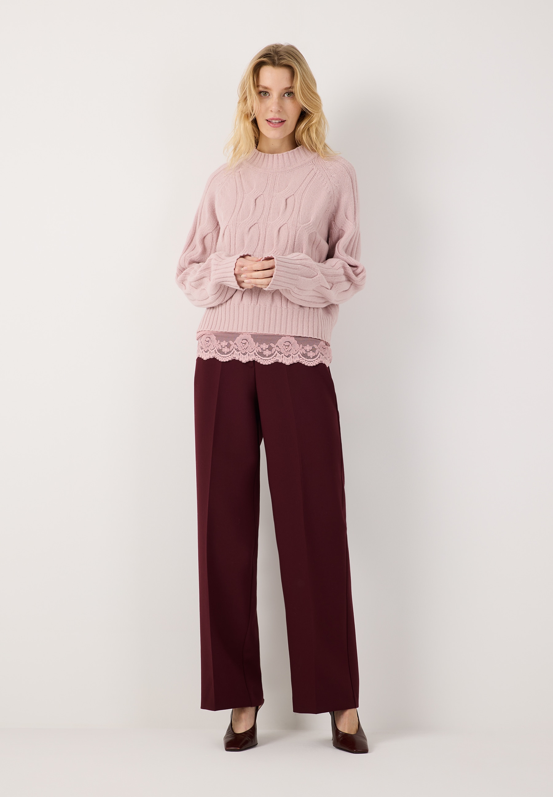 MORE & MORE Pullover in Zopfstrick, rosa, mit weinroter Hose und rosa Spitzen-Top. Model lächelt. MORE & MORE Pullover in Zopfstrick, rosa, mit weinroter Hose und rosa Spitzen-Top. Model lächelt.
