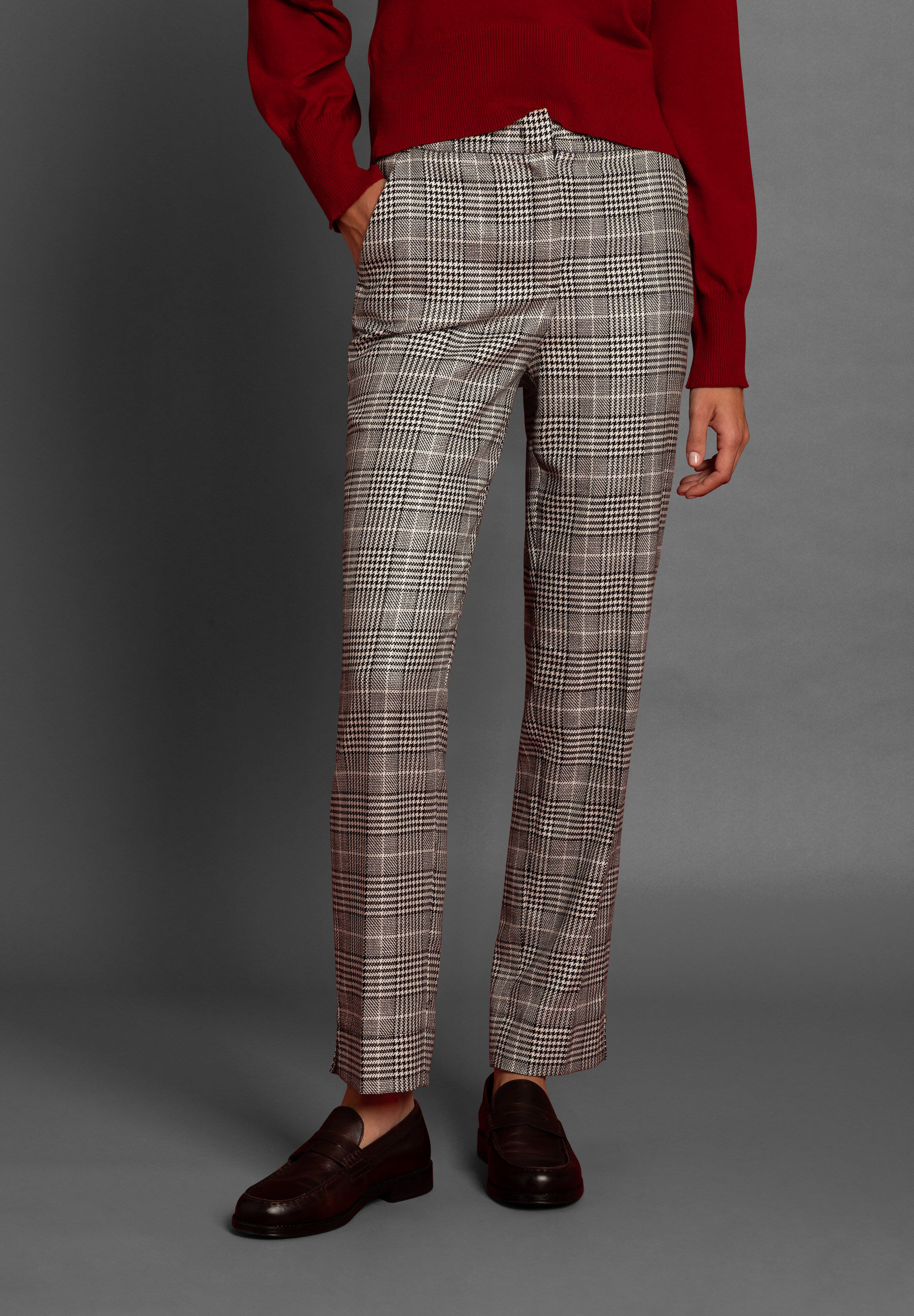 Eine Frau trägt eine karierte Hose von MORE & MORE, kombiniert mit einem roten Oberteil und braunen Schuhen vor grauem Hintergrund.