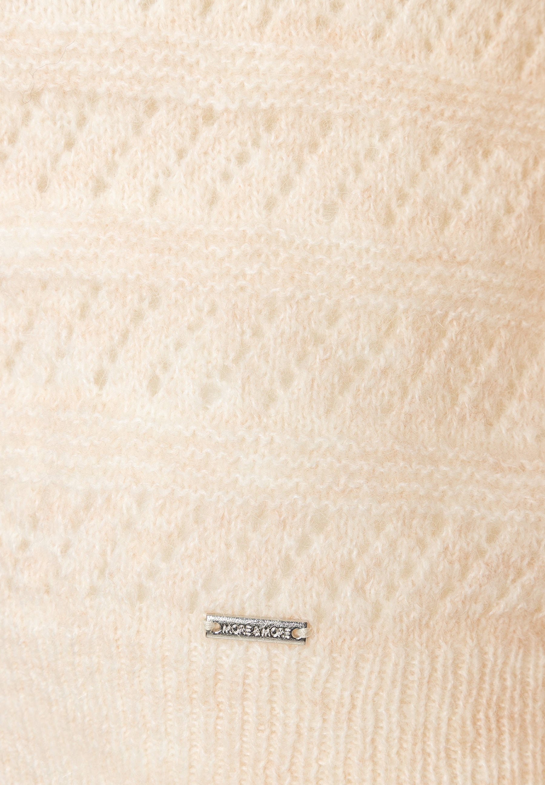 MORE & MORE Pullover. Nahaufnahme beiger Strickware, die ein abstraktes Muster zeigt, mit einem silbernen Logo unten. MORE & MORE Pullover. Nahaufnahme beiger Strickware, die ein abstraktes Muster zeigt, mit einem silbernen Logo unten.