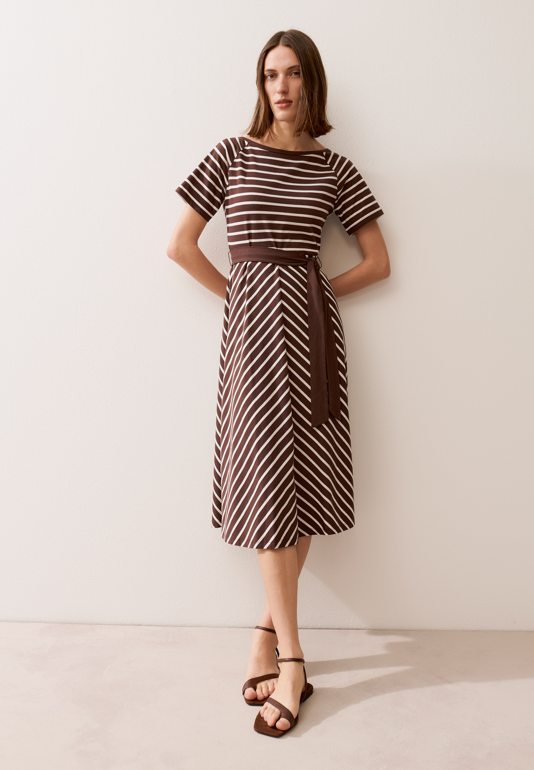 Eine Frau präsentiert ein braun-weiß gestreiftes MORE & MORE Kleid mit Gürtel und braune Sandalen vor einem hellen Hintergrund.