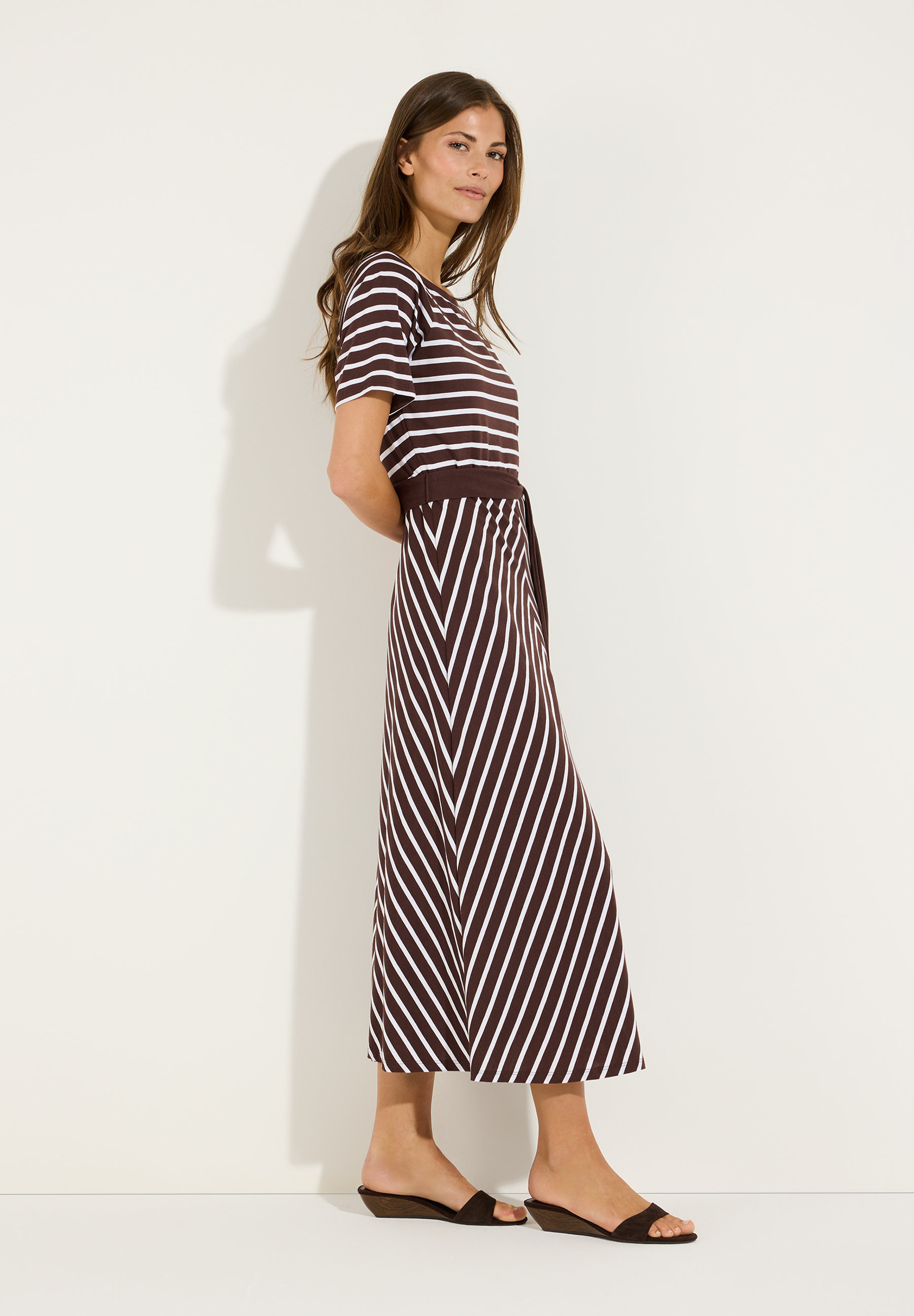 MORE & MORE Jersey-Kleid gestreift. Modell trägt ein braunes Kleid mit weißen Querstreifen oben und diagonalen Streifen unten, dazu braune Sandalen.