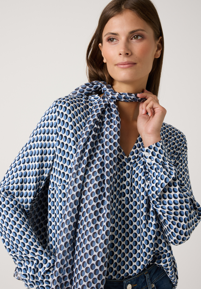 Eine Frau trägt ein MORE & MORE Accessoire, eine blau-weiß gepunktete Bluse mit Kragen. Sie berührt den Kragen mit der Hand. Eine Frau trägt ein MORE & MORE Accessoire, eine blau-weiß gepunktete Bluse mit Kragen. Sie berührt den Kragen mit der Hand.