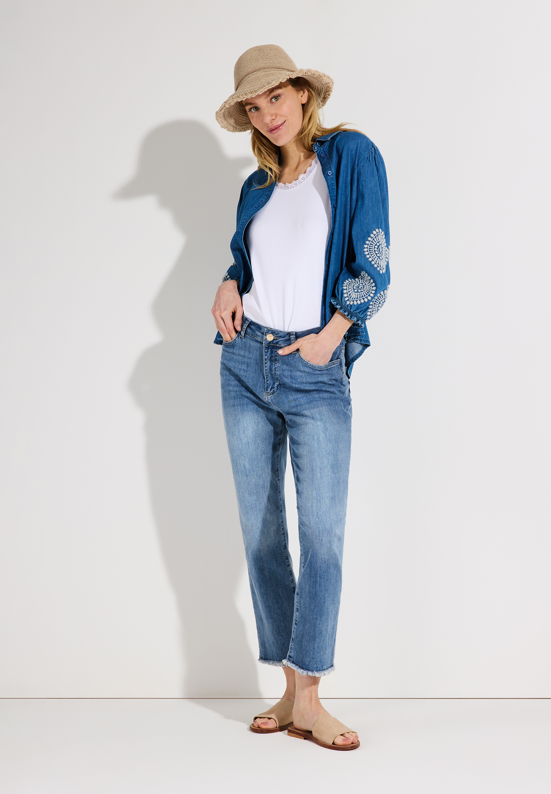 Eine Frau mit MORE & MORE Hut, Jeansjacke und Jeans posiert vor einem weißen Hintergrund. Eine Frau mit MORE & MORE Hut, Jeansjacke und Jeans posiert vor einem weißen Hintergrund.