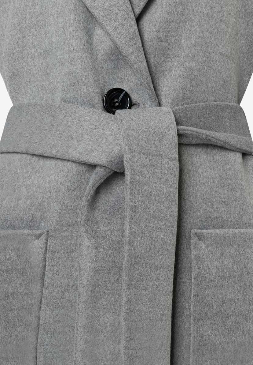 Nahaufnahme eines grauen MORE & MORE Oberteils mit Bindegürtel und Knopfdetail. Nahaufnahme eines grauen MORE & MORE Oberteils mit Bindegürtel und Knopfdetail.