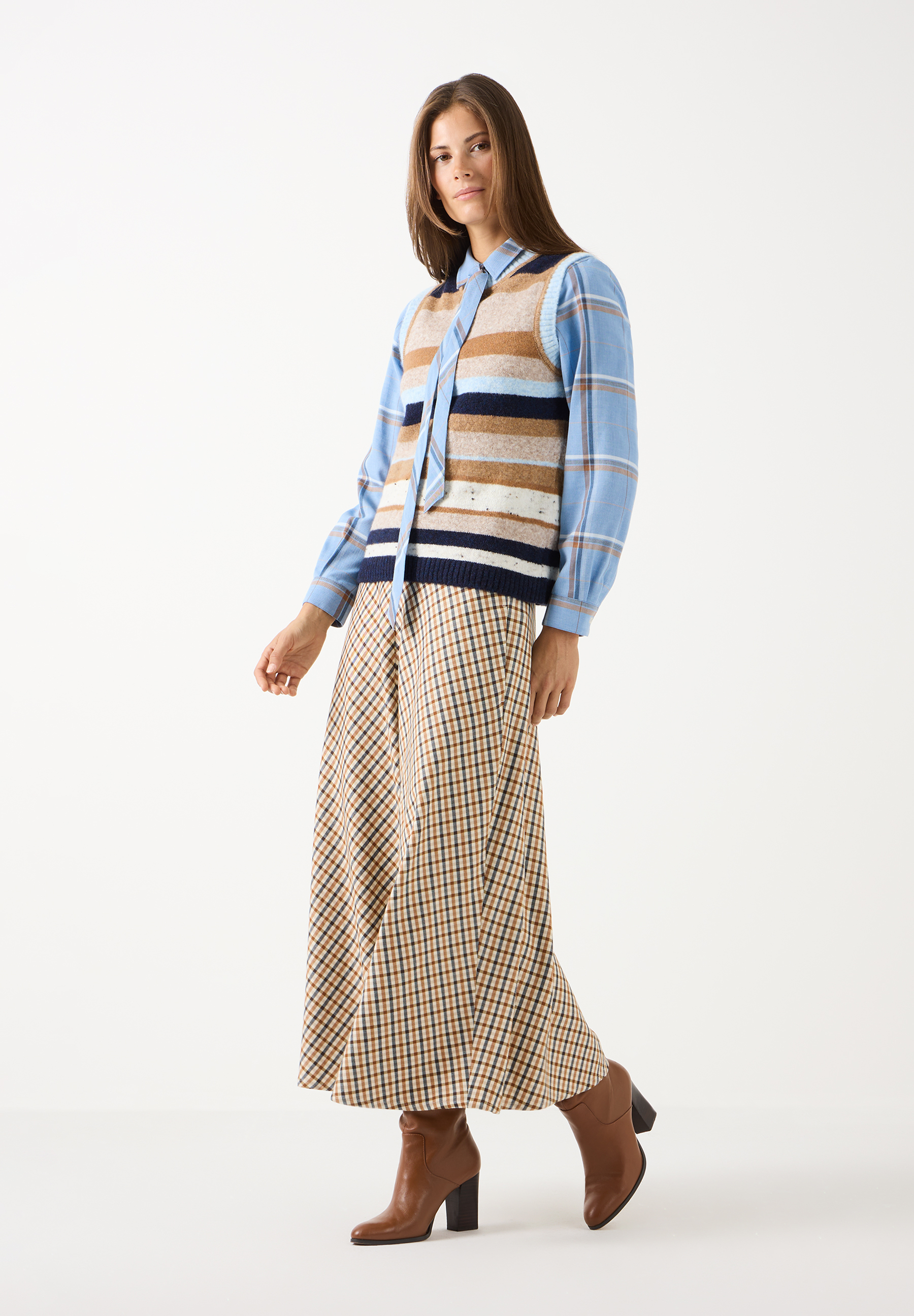 MORE & MORE Bluse: Eine Frau trägt eine blau karierte MORE & MORE Bluse unter einem gestreiften Strickpullunder in Blau- und Brauntönen. Dazu einen karierten Rock und braune Stiefeletten.
