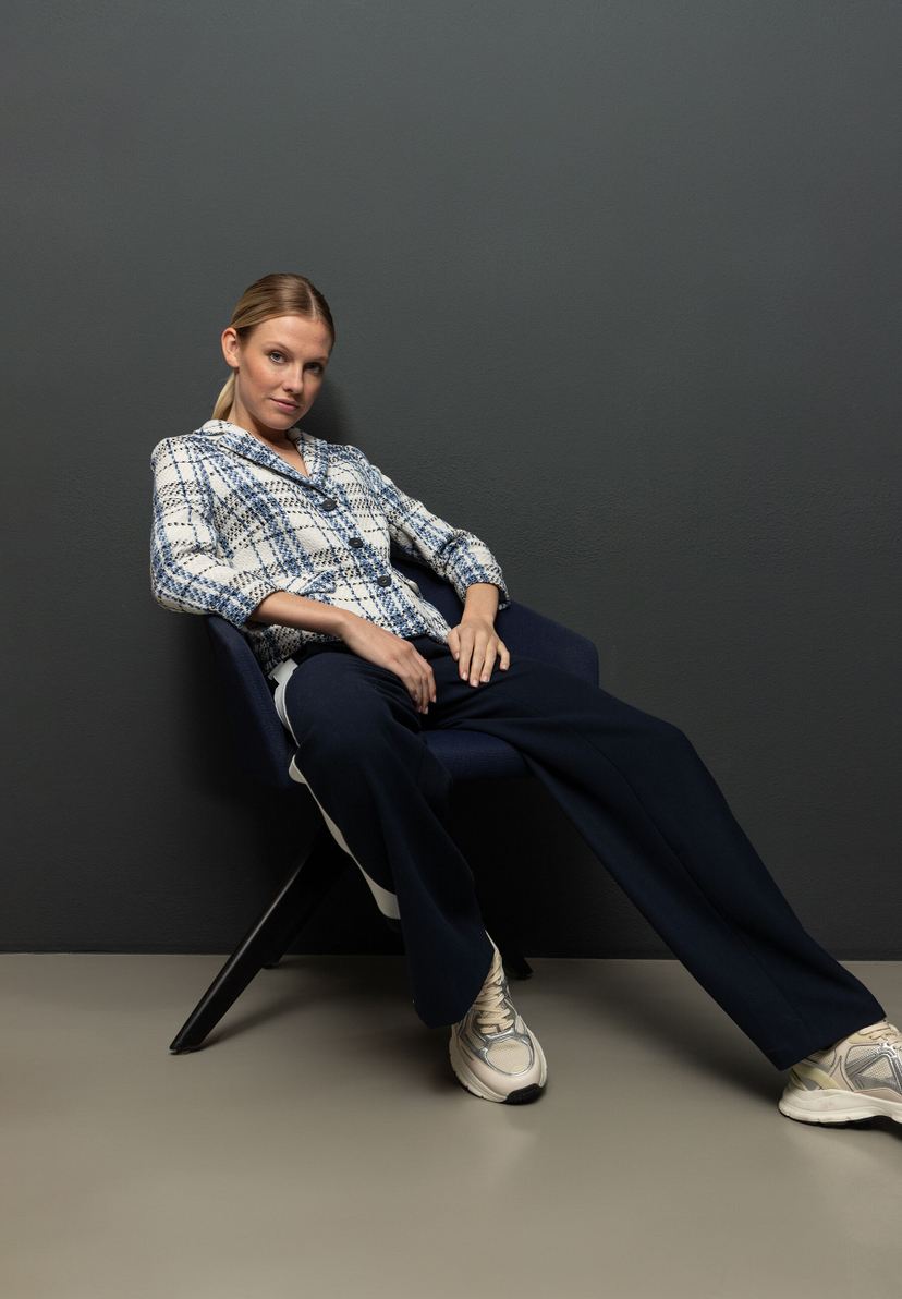 Eine Frau in einem Bouclé-Blazer von MORE & MORE sitzt auf einem Stuhl vor einem grauen Hintergrund. Eine Frau in einem Bouclé-Blazer von MORE & MORE sitzt auf einem Stuhl vor einem grauen Hintergrund.