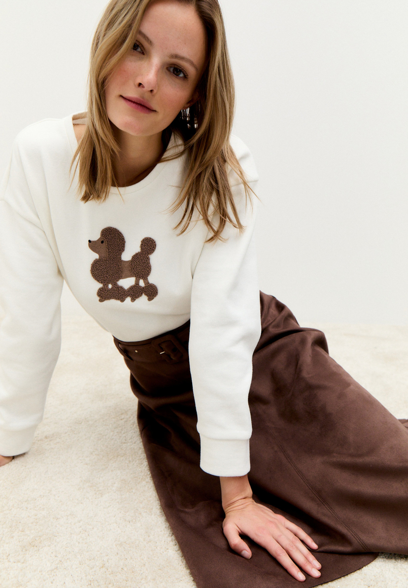 Eine Frau sitzt im MORE & MORE-Sweatshirt mit Pudel-Applikation und braunem Rock auf einem hellen Teppich. Eine Frau sitzt im MORE & MORE-Sweatshirt mit Pudel-Applikation und braunem Rock auf einem hellen Teppich.