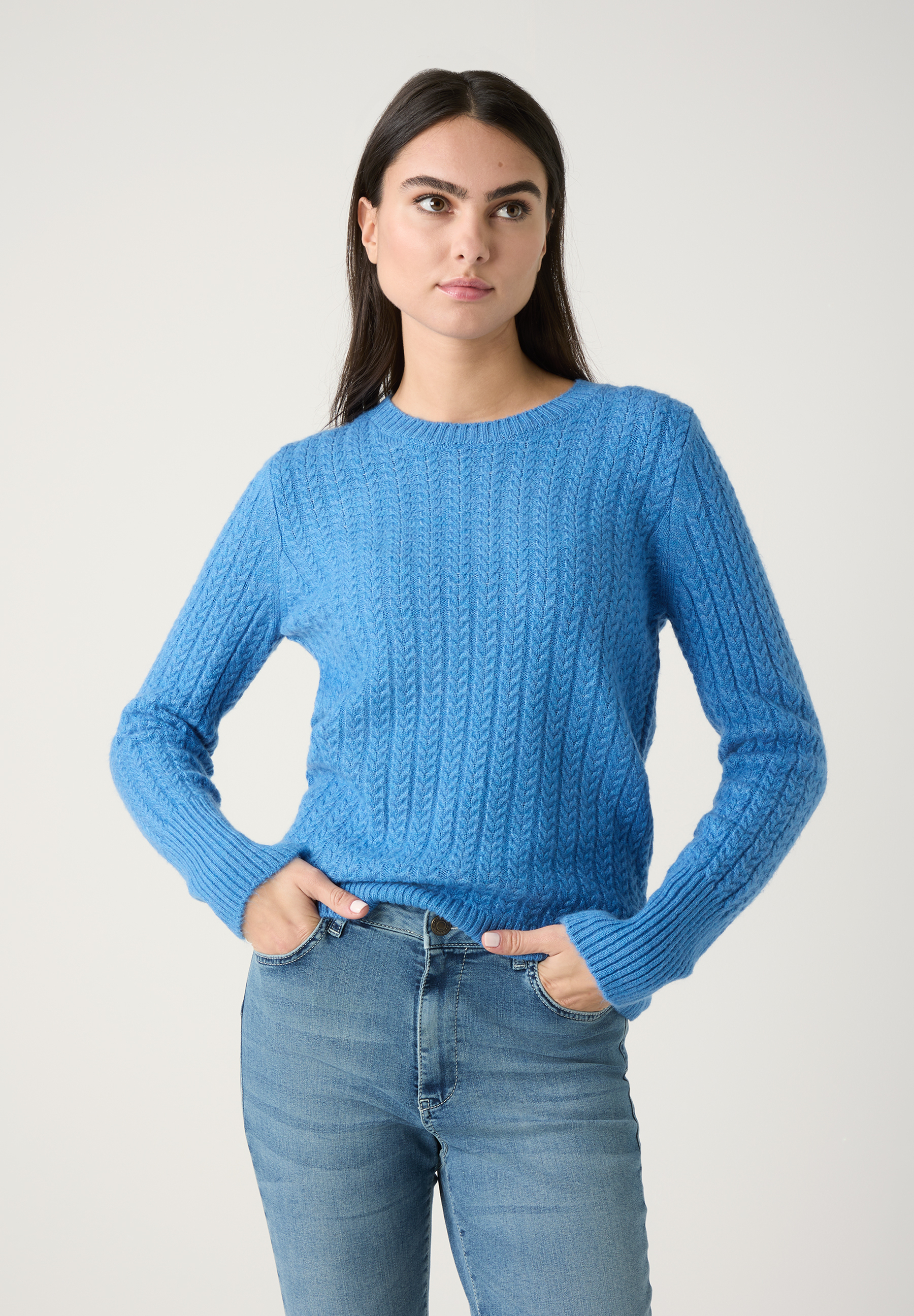Eine Frau mit MORE & MORE Strickpullover in Blau und Jeans, Hände in den Hosentaschen.