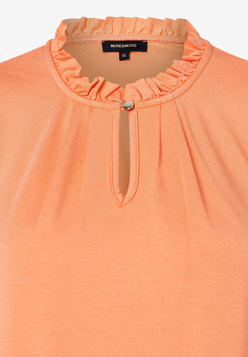 Nahaufnahme eines pfirsichfarbenen Jersey-Oberteils von MORE & MORE mit Rüschenkragen und Knopfdetail. Nahaufnahme eines pfirsichfarbenen Jersey-Oberteils von MORE & MORE mit Rüschenkragen und Knopfdetail.