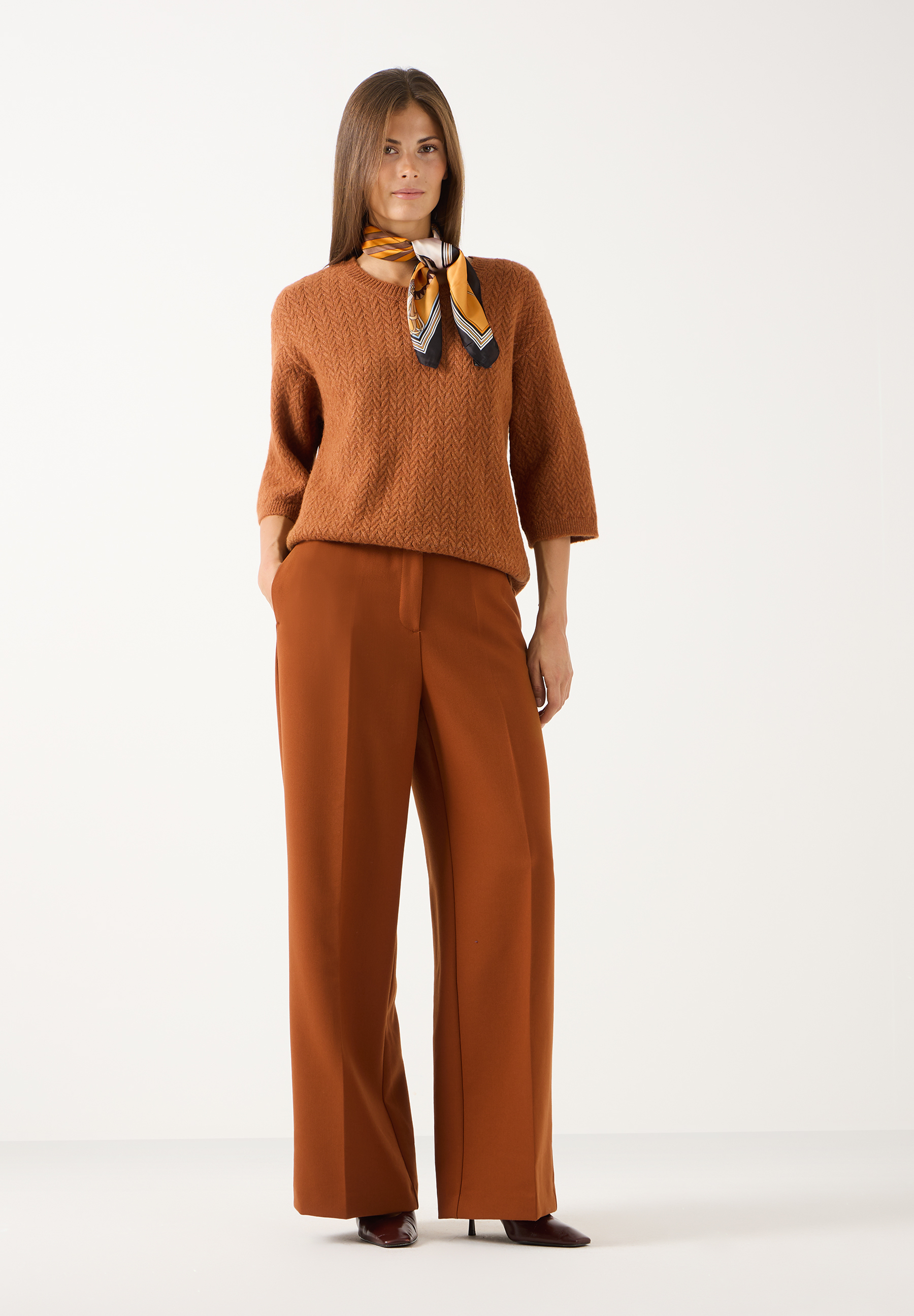 MORE & MORE Accessoires: Eine Frau mit einem mehrfarbigen Seidentuch, braunem Pullover und passenden Stoffhosen.