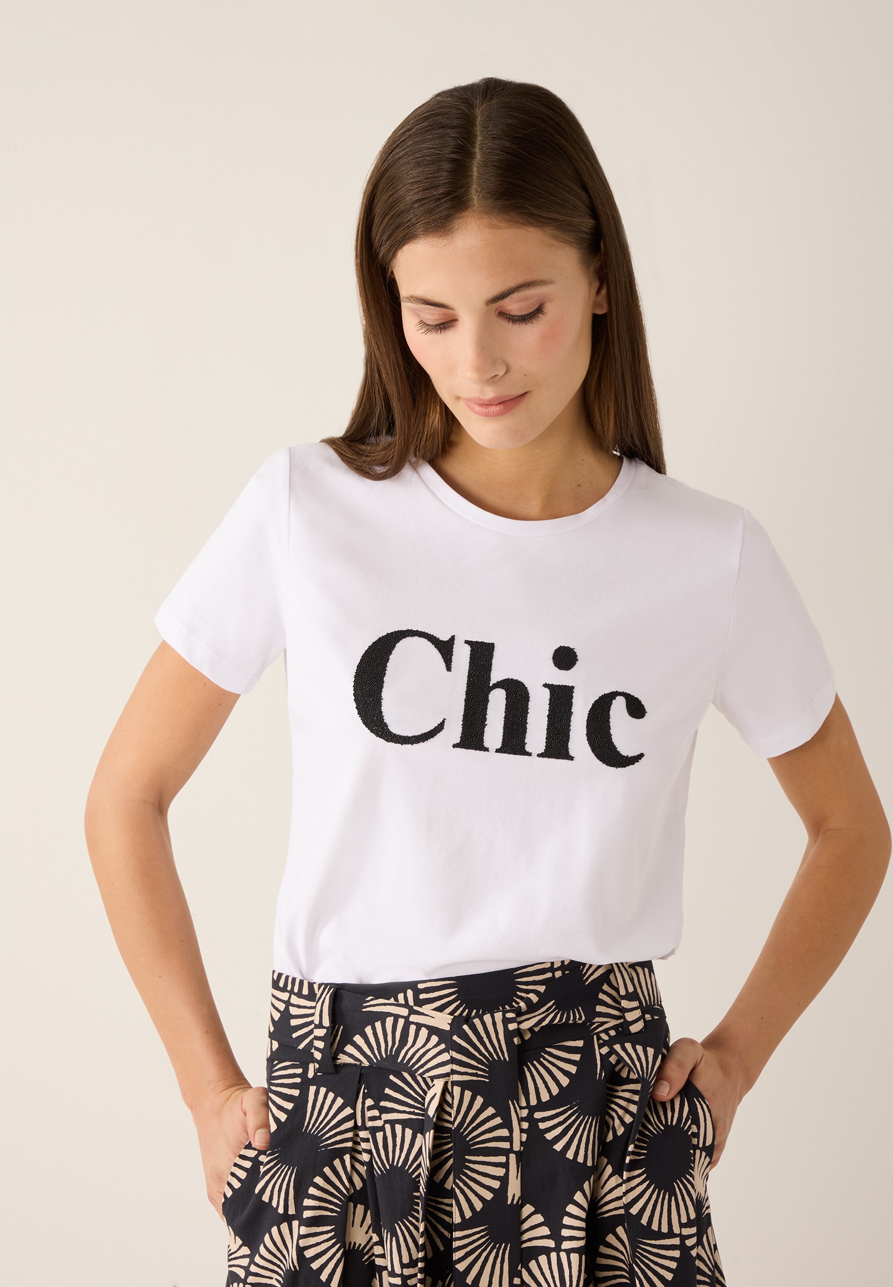 Eine Frau trägt ein MORE & MORE T-Shirt mit dem Wort "Chic" und eine gemusterte Hose.