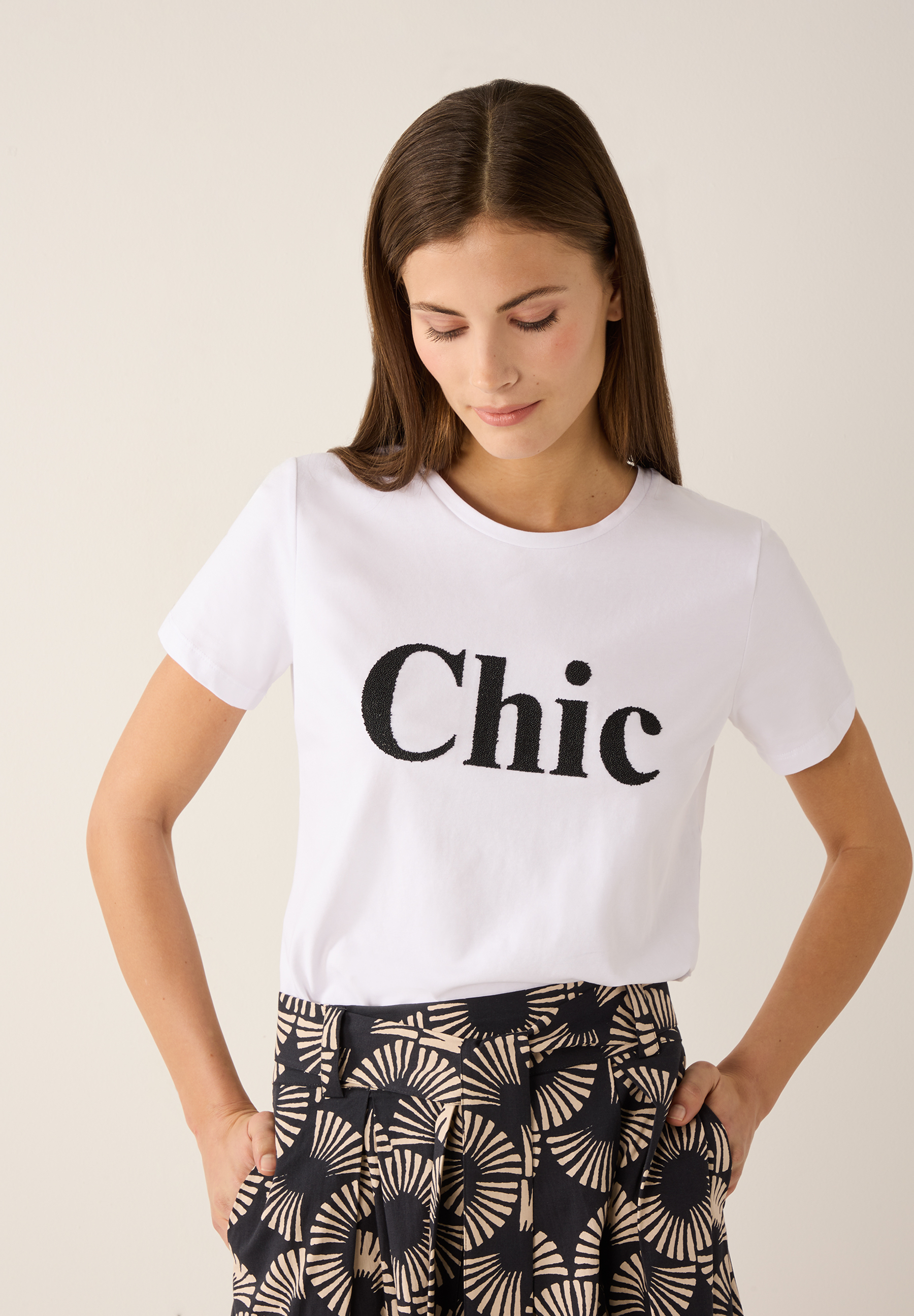 Eine Frau trägt ein MORE & MORE T-Shirt mit dem Wort "Chic" und eine gemusterte Hose.