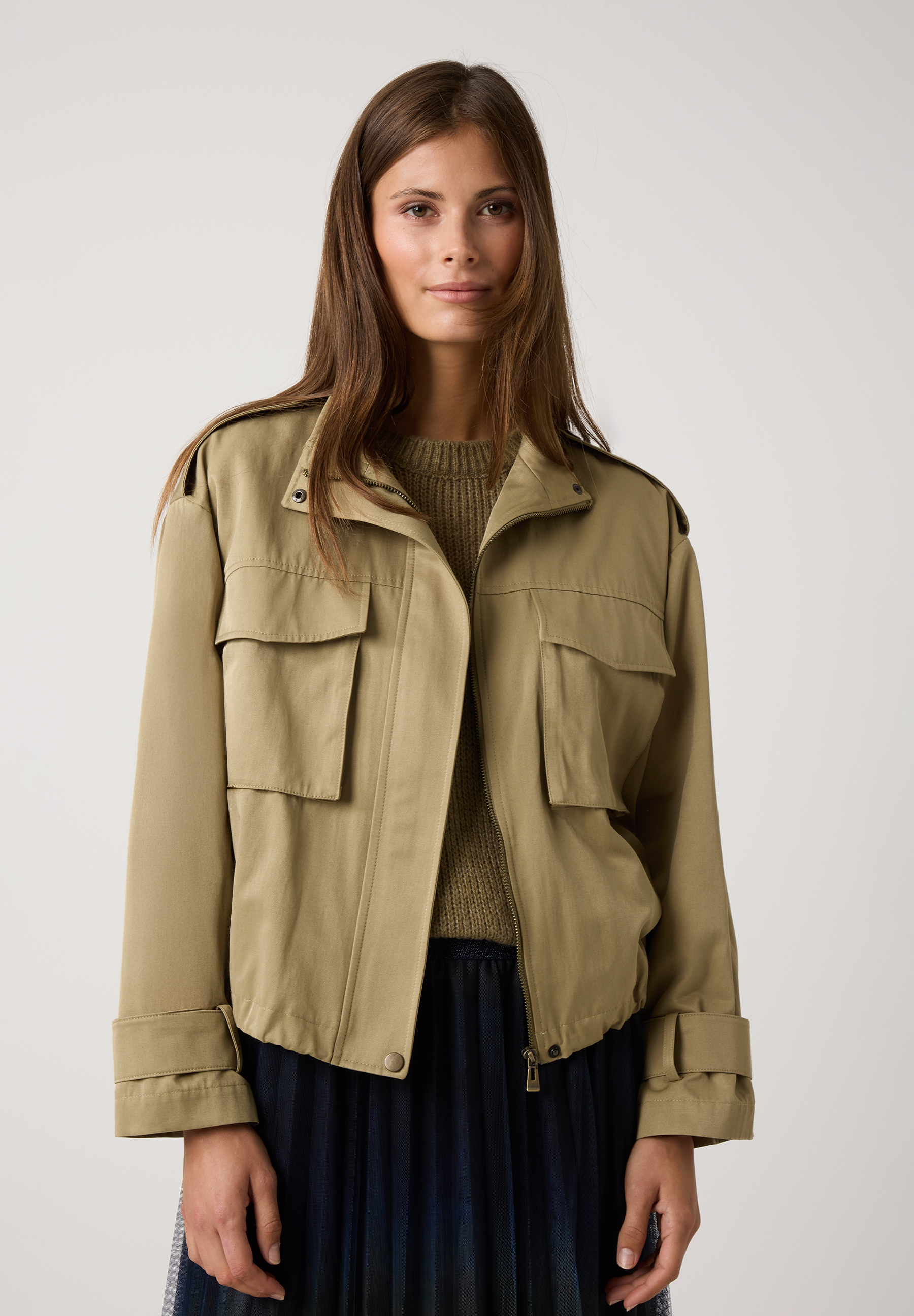 Eine Frau trägt eine Khaki-Jacke von MORE & MORE. Die Jacke hat aufgesetzte Taschen und Schulterklappen.